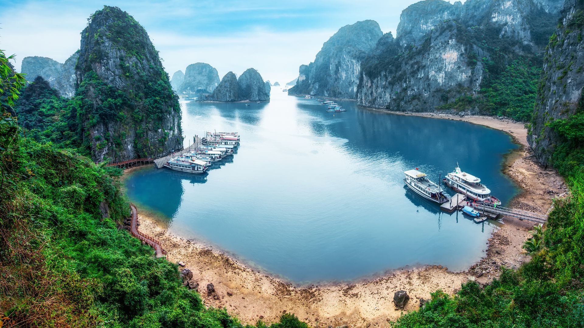 An aerial photograph showing many traditional Vietnamese tour boats and cruise ships in the clear, blue-green waters of Hạ Long Bay, surrounded by towering, jungle-covered limestone islands under a bright sky.