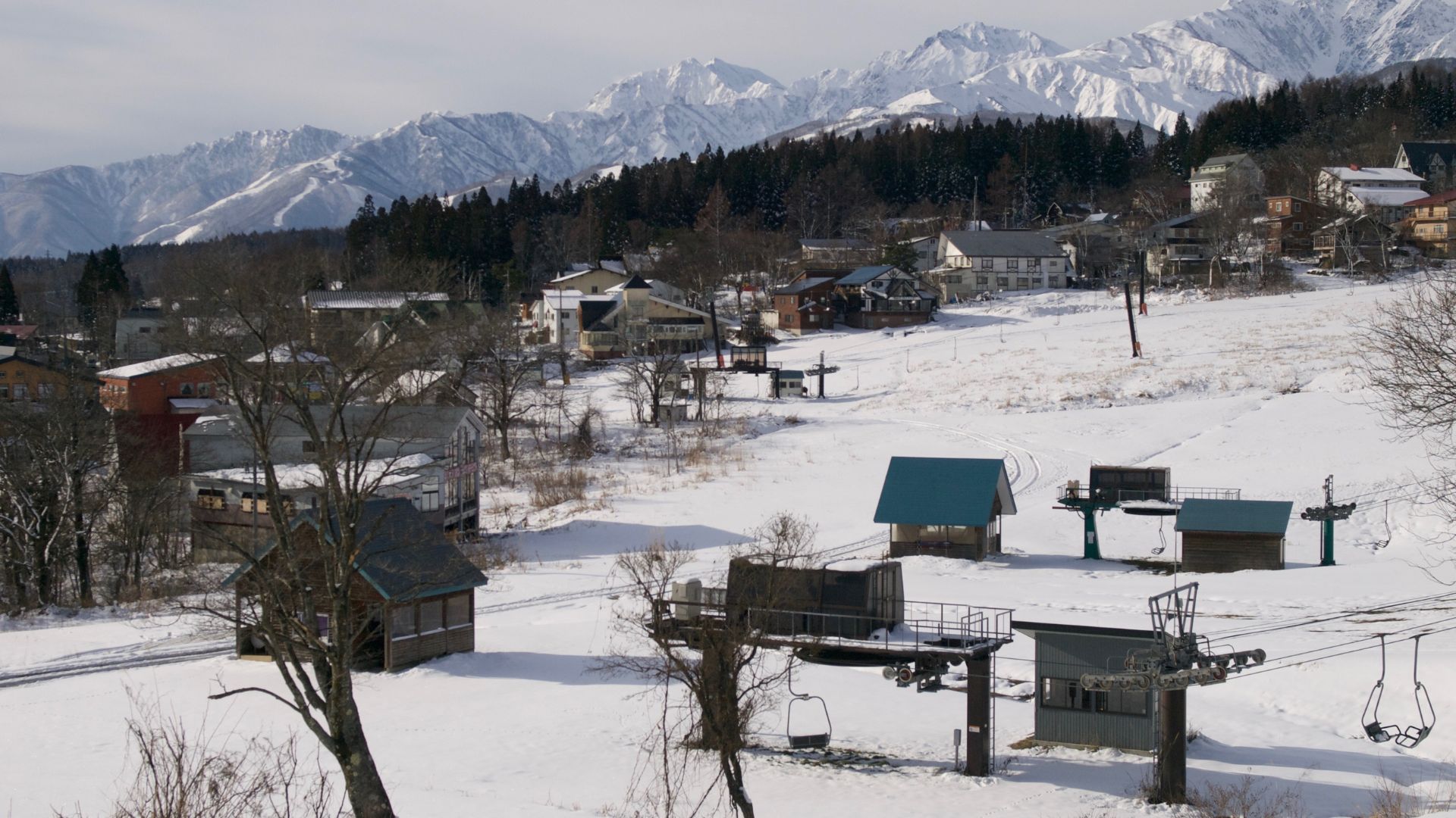 A snow-covered ski resort village with buildings and dormant chairlifts sits in the foreground, with large, snow-capped mountains of the Japanese Alps rising prominently in the background under a pale sky.