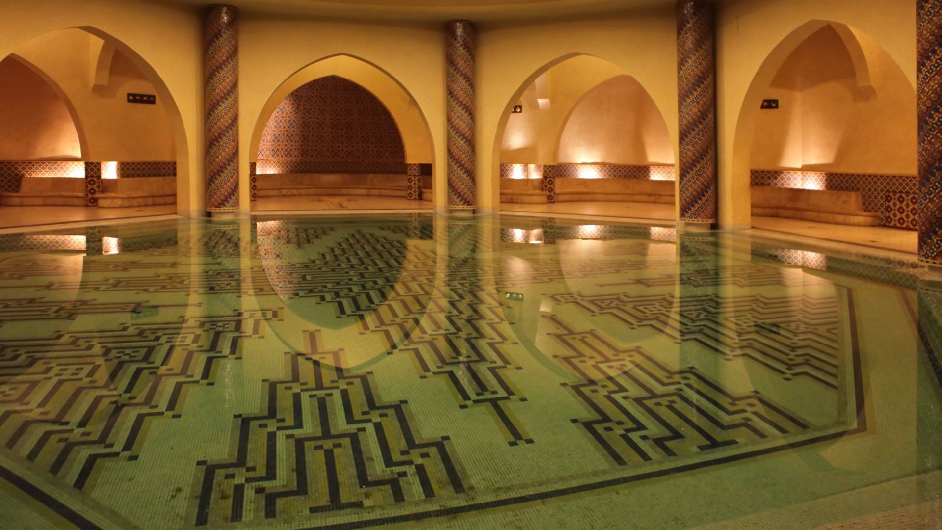 A bathing pool inside the Hammam of the Hassan II Mosque in Casablanca, Morocco, featuring arched doorways and a floor with reflective green water and dark geometric mosaic patterns.