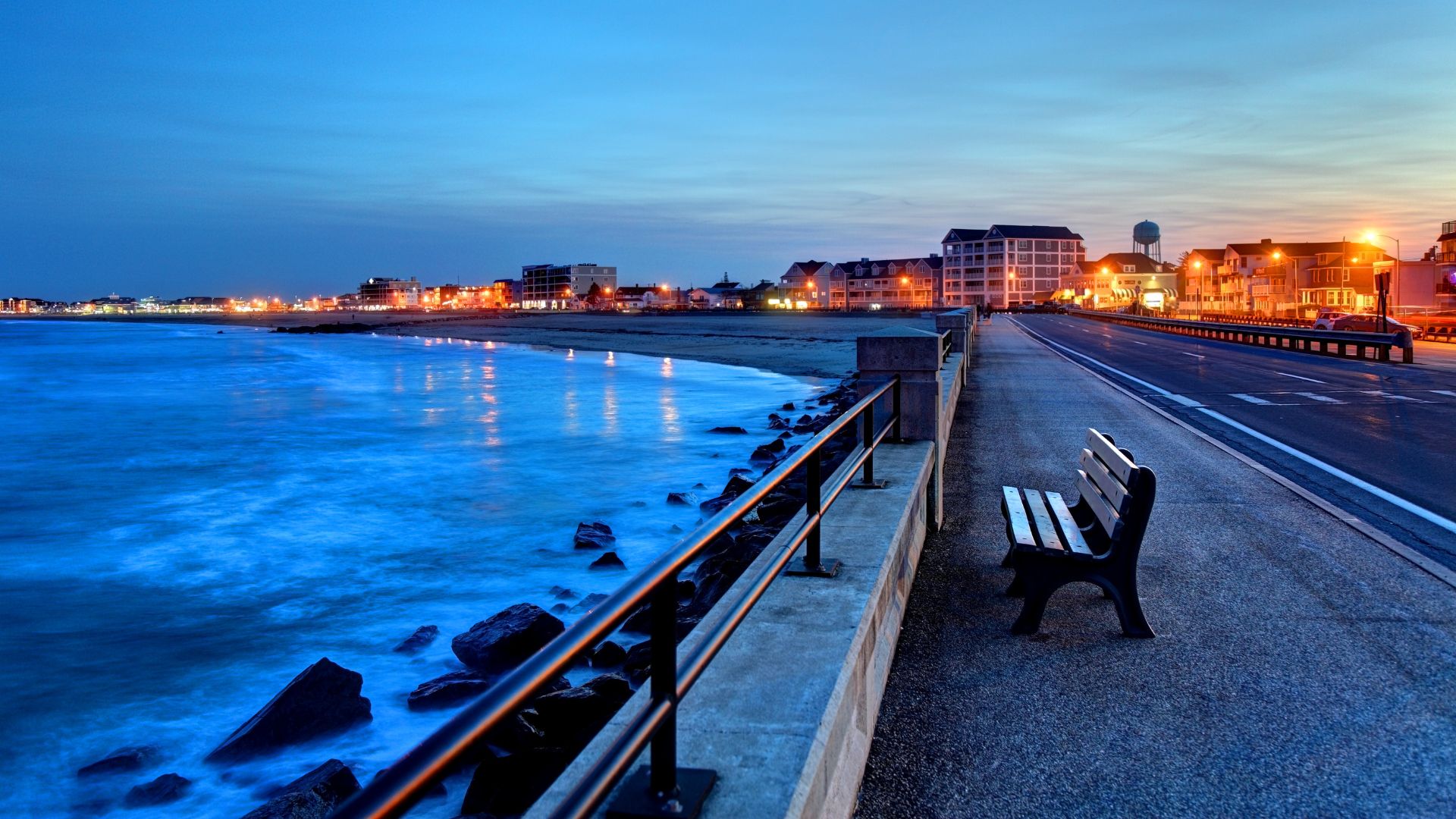 A coastal scene at dusk with a dark sky, a paved walkway, a metal railing, and ocean waves crashing against rocks in the foreground, leading to a developed beachfront town skyline in the distance.