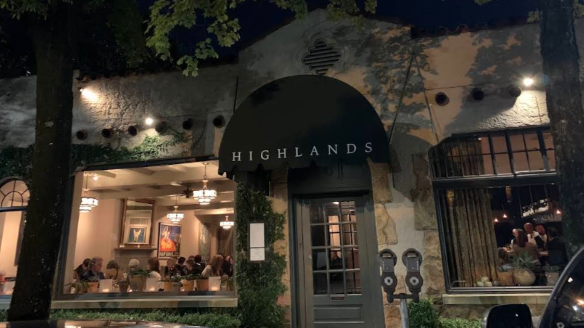 An evening photo of the exterior of Highlands Bar & Grill in Birmingham, Alabama, showing the ivy-covered stone facade, a prominent dark awning over the entrance with the restaurant's name, and warmly lit dining areas visible through the windows.