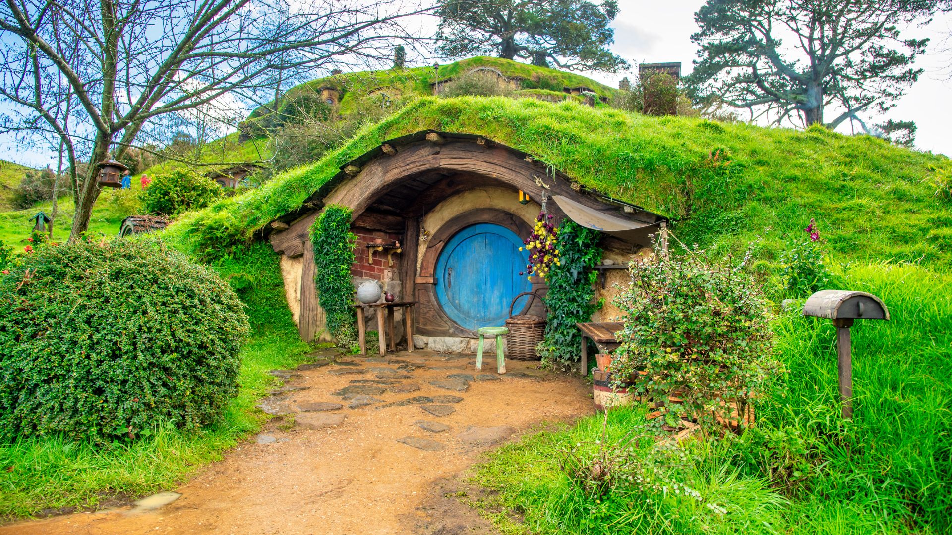 A round blue door, belonging to a house built into a lush green hill, sits at the end of a dirt path.
