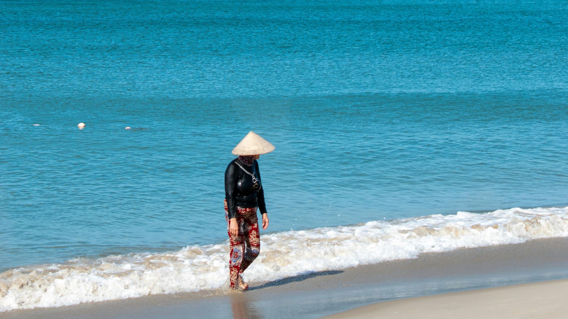 A person wearing a traditional conical hat walks along the white-capped ocean shore in Vietnam, under a blue sky.