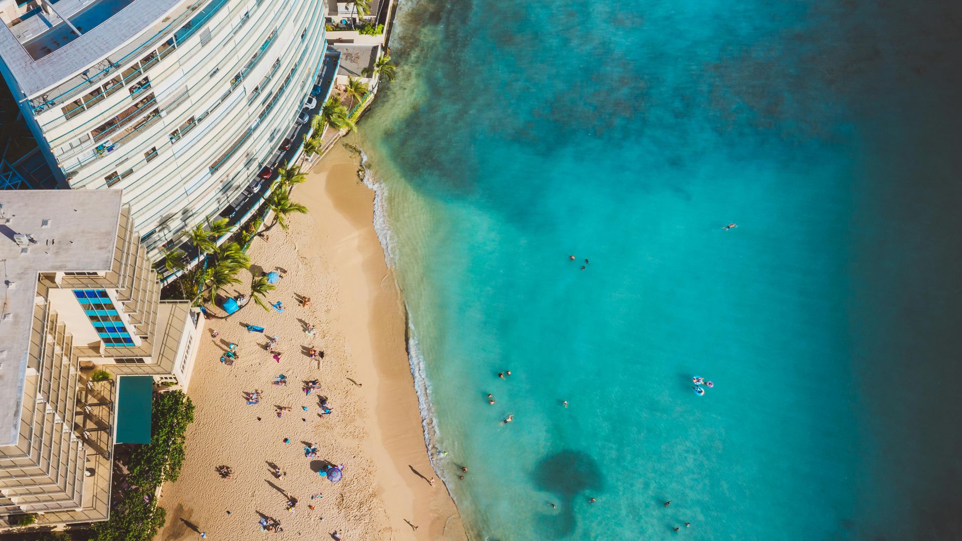 An aerial view of a busy sandy beach next to a large oceanfront hotel, with people swimming in the clear turquoise water.