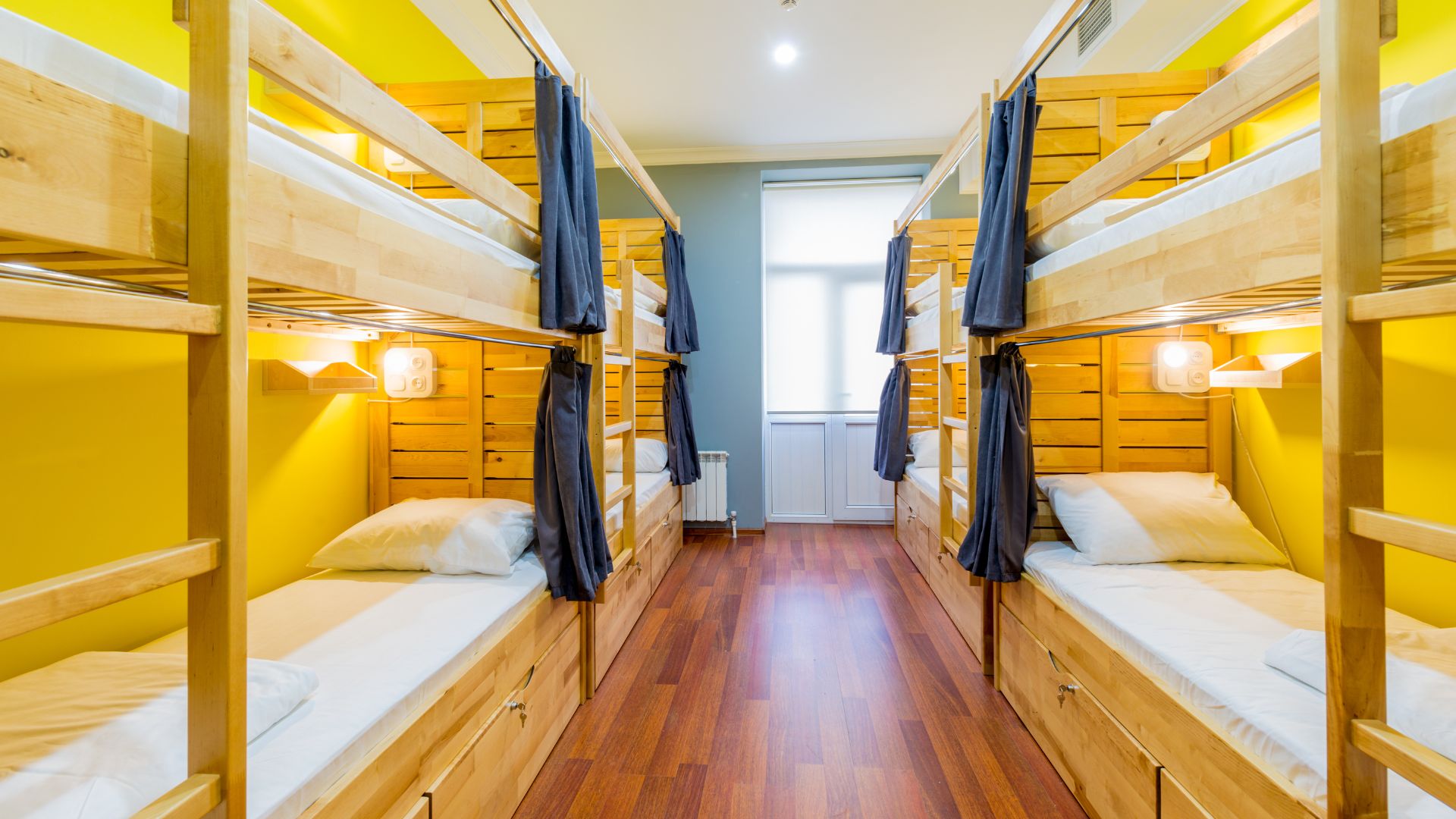An empty hostel dorm room with wooden bunk beds, yellow walls, a wood floor, and privacy curtains.