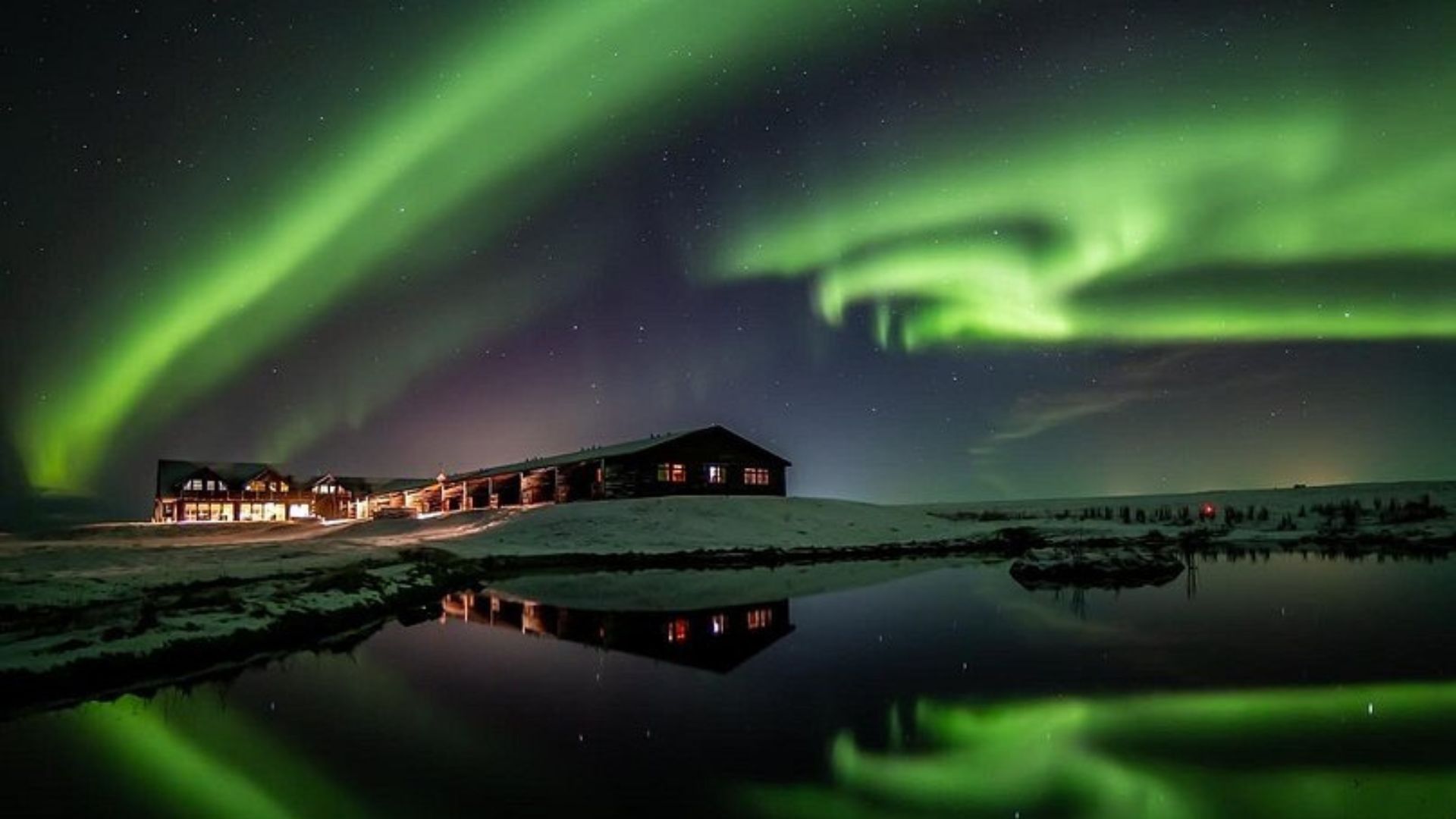 An alpine-style log hotel with warm lights is reflected in a calm pool of water, set against a snow-dusted landscape under a dark night sky filled with vivid green, pink, and purple streaks of the Aurora Borealis.