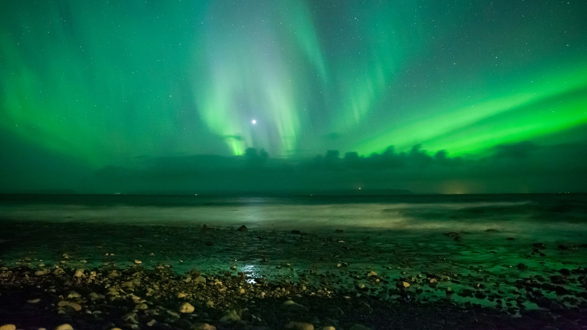 A small, lit coastal village and a dark hill under a night sky filled with a vibrant, dancing green aurora borealis reflecting on the water in the foreground.