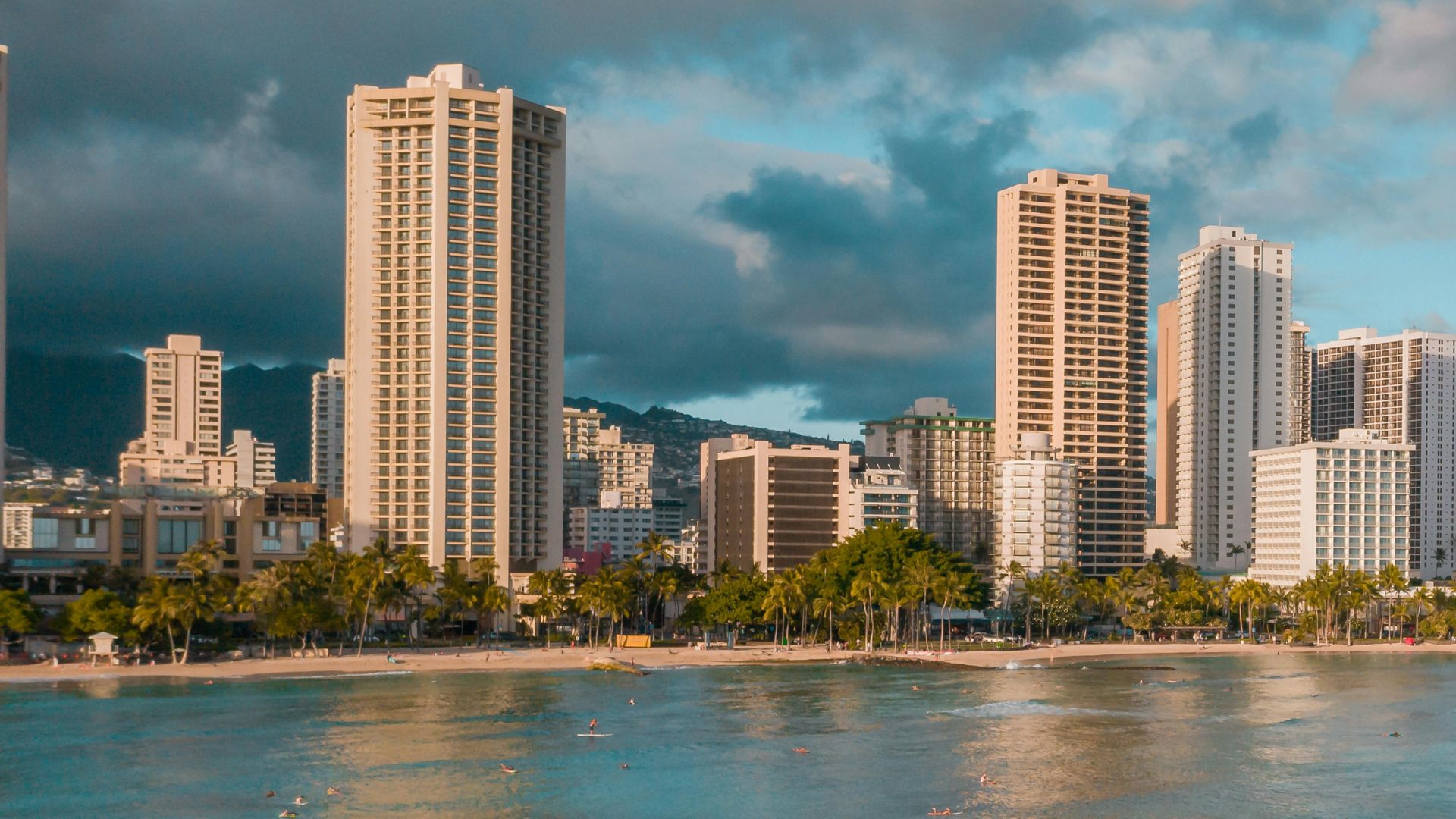 A sunny day view of a crowded Waikīkī beach with turquoise ocean waters, palm trees, modern high-rise resort hotels lining the shore, and the iconic Diamond Head crater visible in the background under a blue sky with white clouds.