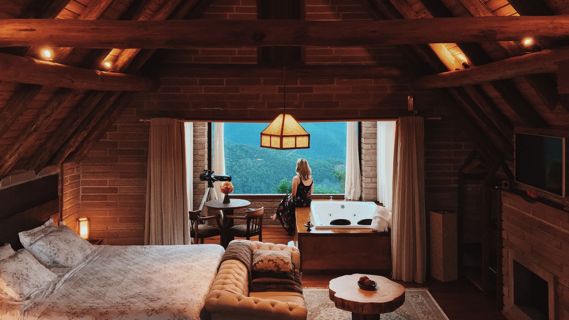 A cozy, rustic attic hotel room with wooden beams and a large window, where a woman sits looking out at a view of lush green mountainsides.