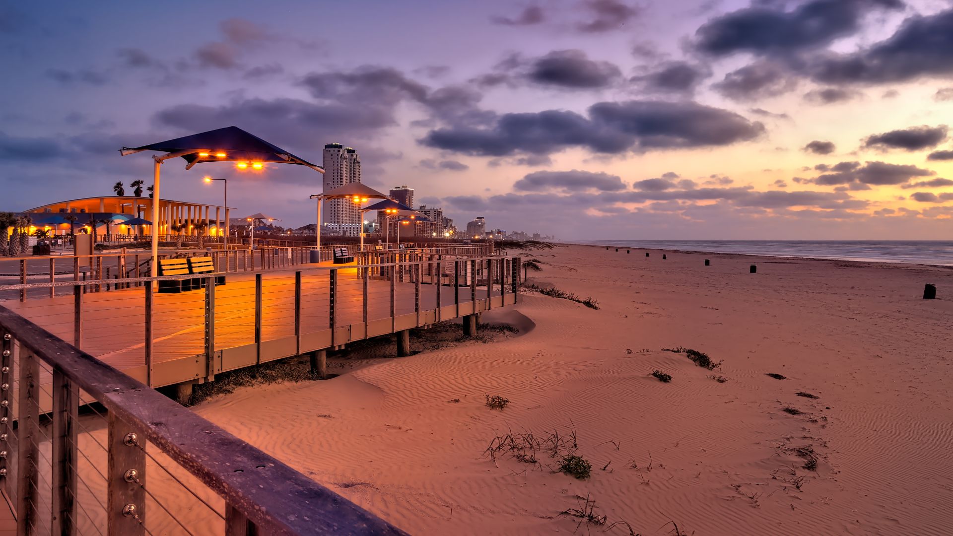 A view along the illuminated wooden boardwalk and sandy beach at Isla Blanca Park, South Padre Island, at sunset or dusk, with buildings and the ocean in the background.