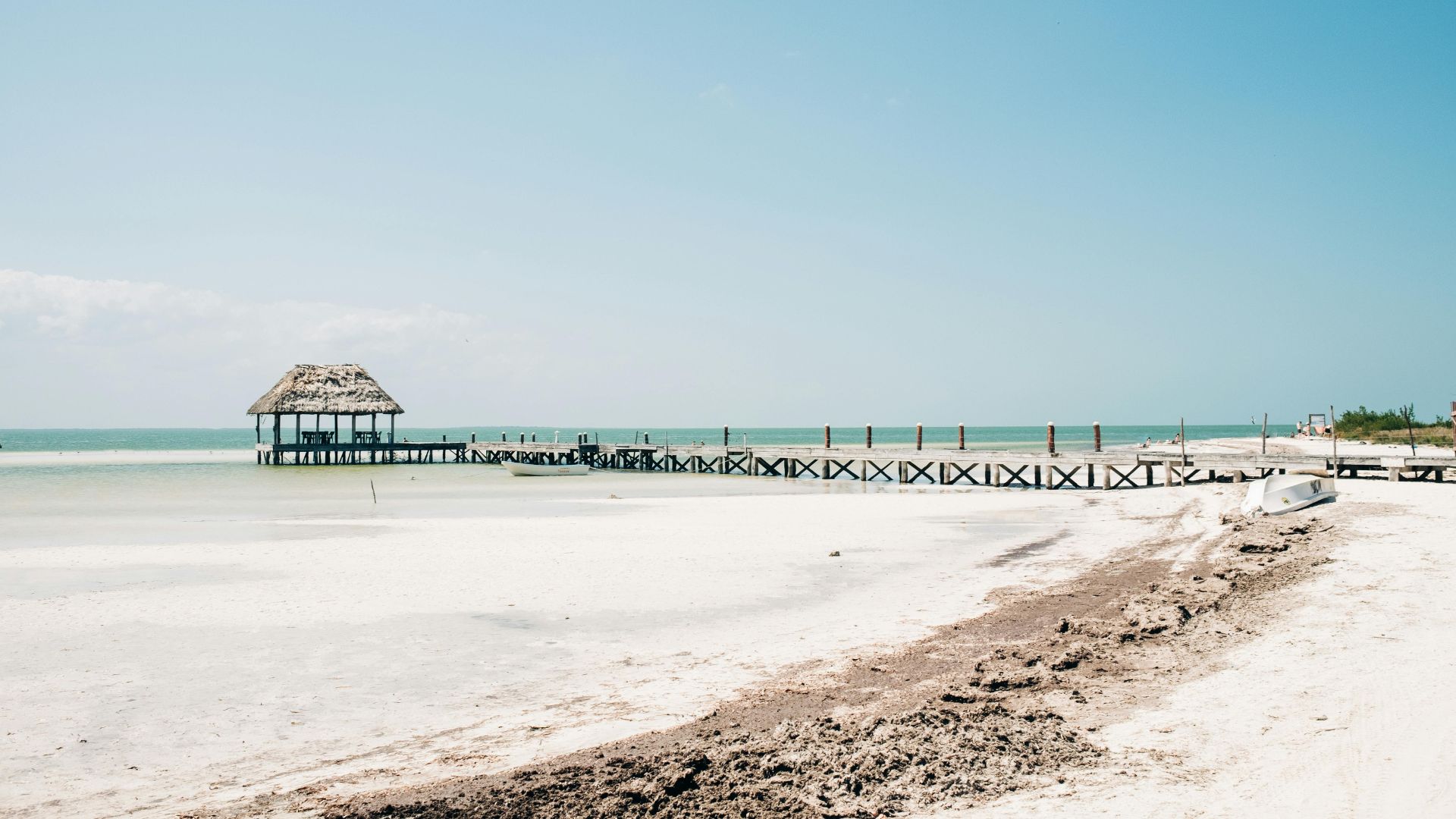 A wide, white-sand beach featuring a long wooden pier extending into the shallow, turquoise ocean water, which leads to a small, thatched-roof palapa at the end, all under a clear blue sky.