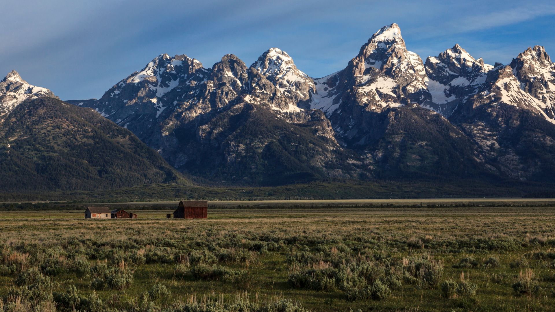 A historic wooden barn sits in a grassy field with the snow-capped, rugged peaks of the Teton Range rising dramatically in the background under a blue sky.