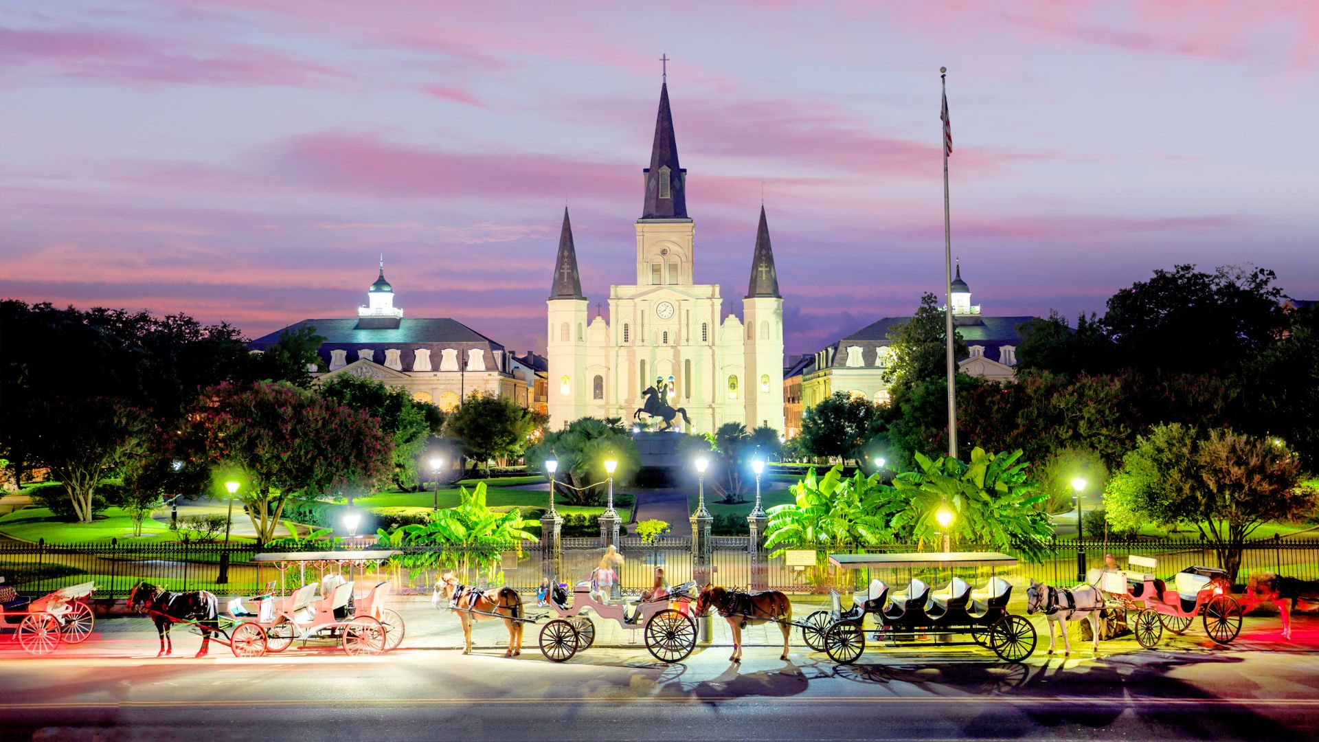 The historic St. Louis Cathedral towering over Jackson Square in the French Quarter of New Orleans at twilight, with several horse-drawn carriages in the illuminated foreground.