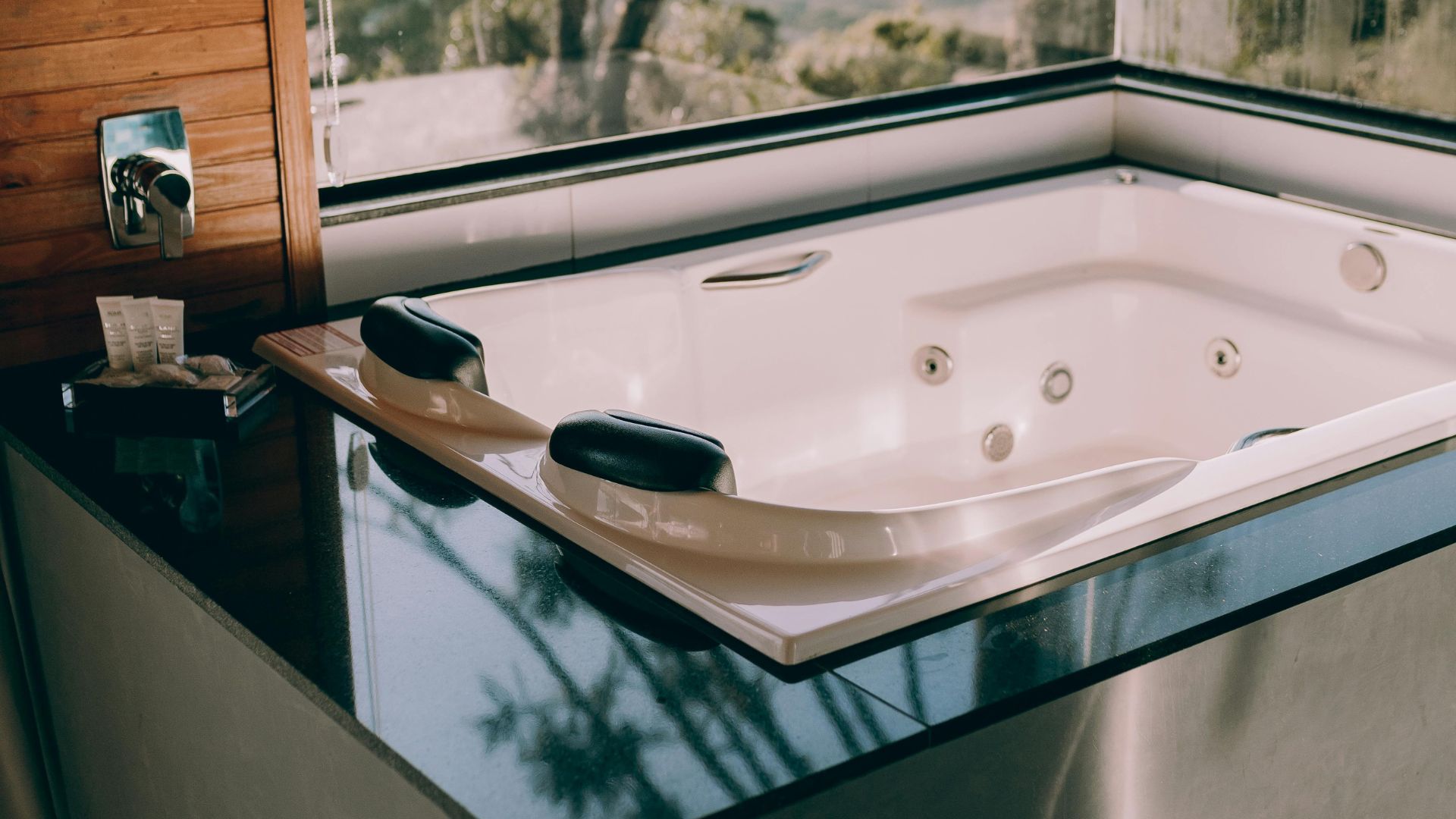 A white built-in jacuzzi tub with black headrests, set into a black polished countertop next to a window overlooking a natural outdoor scene.