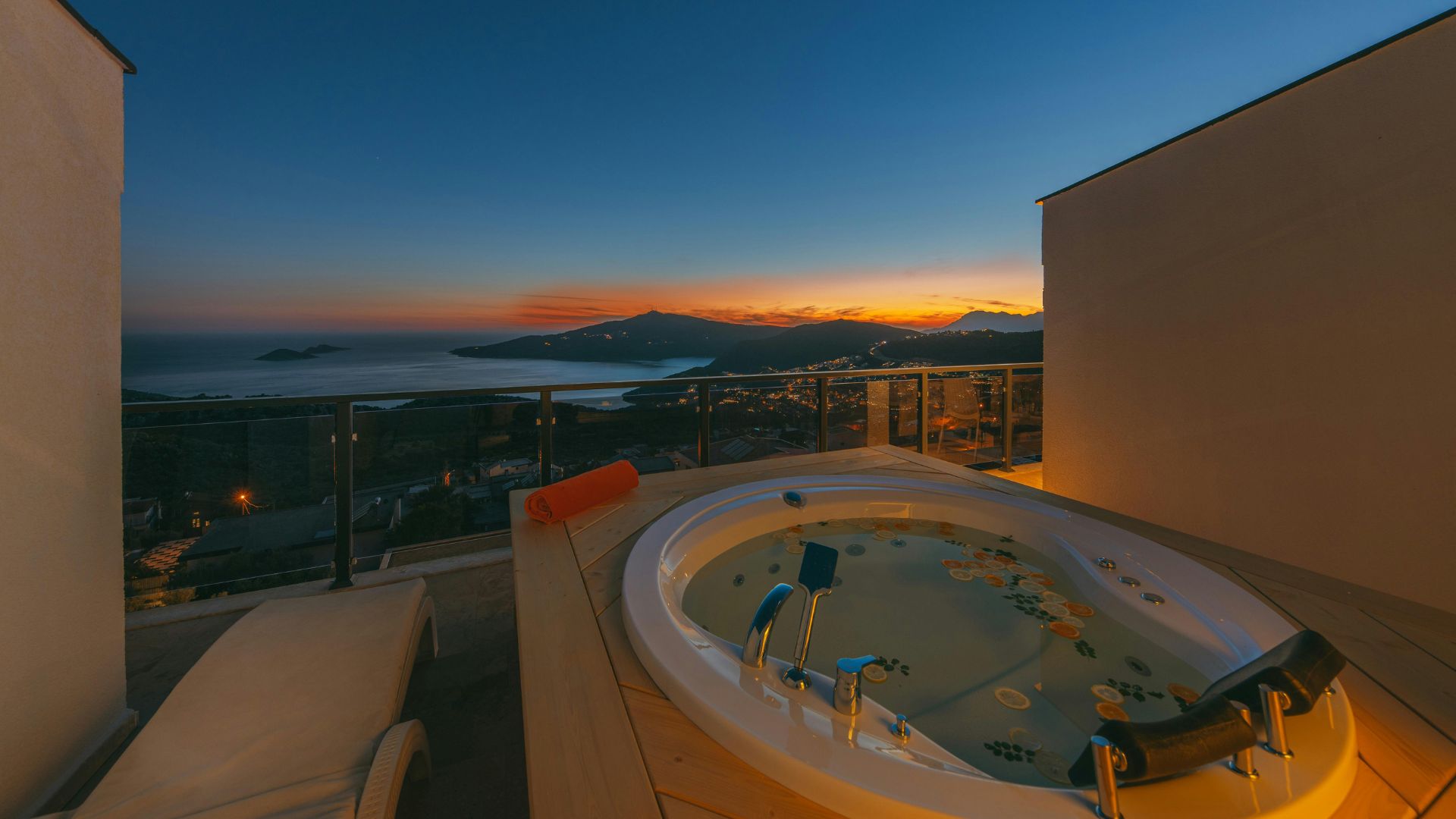 A view from a private balcony featuring an outdoor jacuzzi and lounge chair at dusk, overlooking a coastline with mountains and the sea illuminated by a vibrant orange and yellow sunset sky.