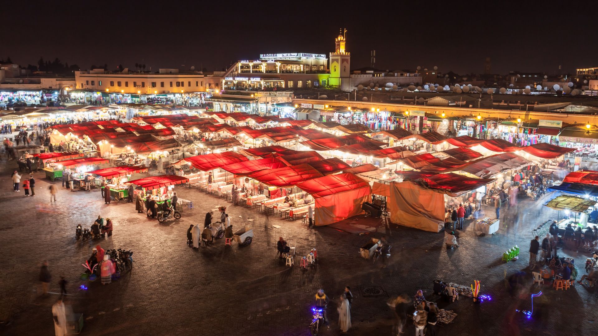 A high-angle view of the bustling Jemaa el-Fnaa market square in Marrakech, Morocco at night, with many stalls featuring red-tented roofs and crowds of people.