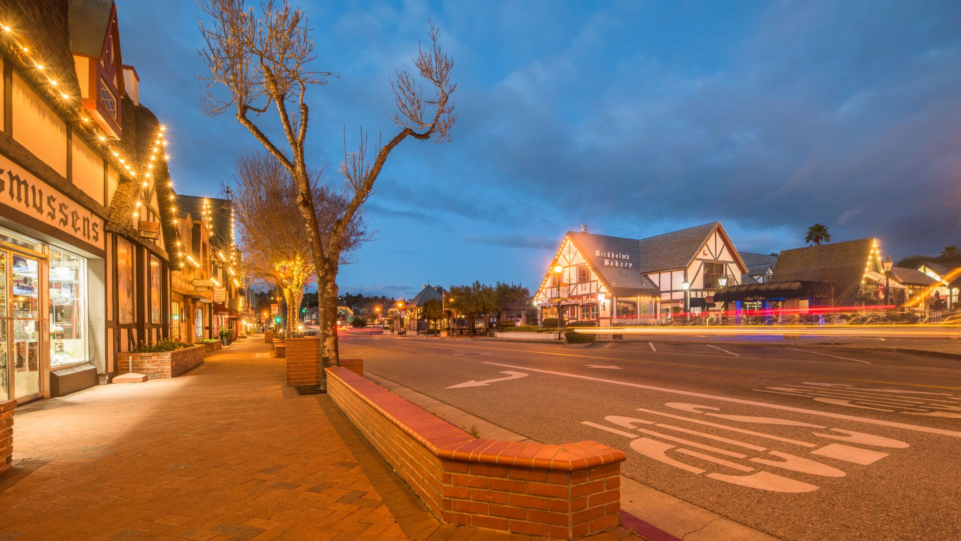 A street in Solvang, California, a town known for its Danish-style architecture, is shown at dusk with holiday lights on the buildings and a long exposure of car lights on the road.