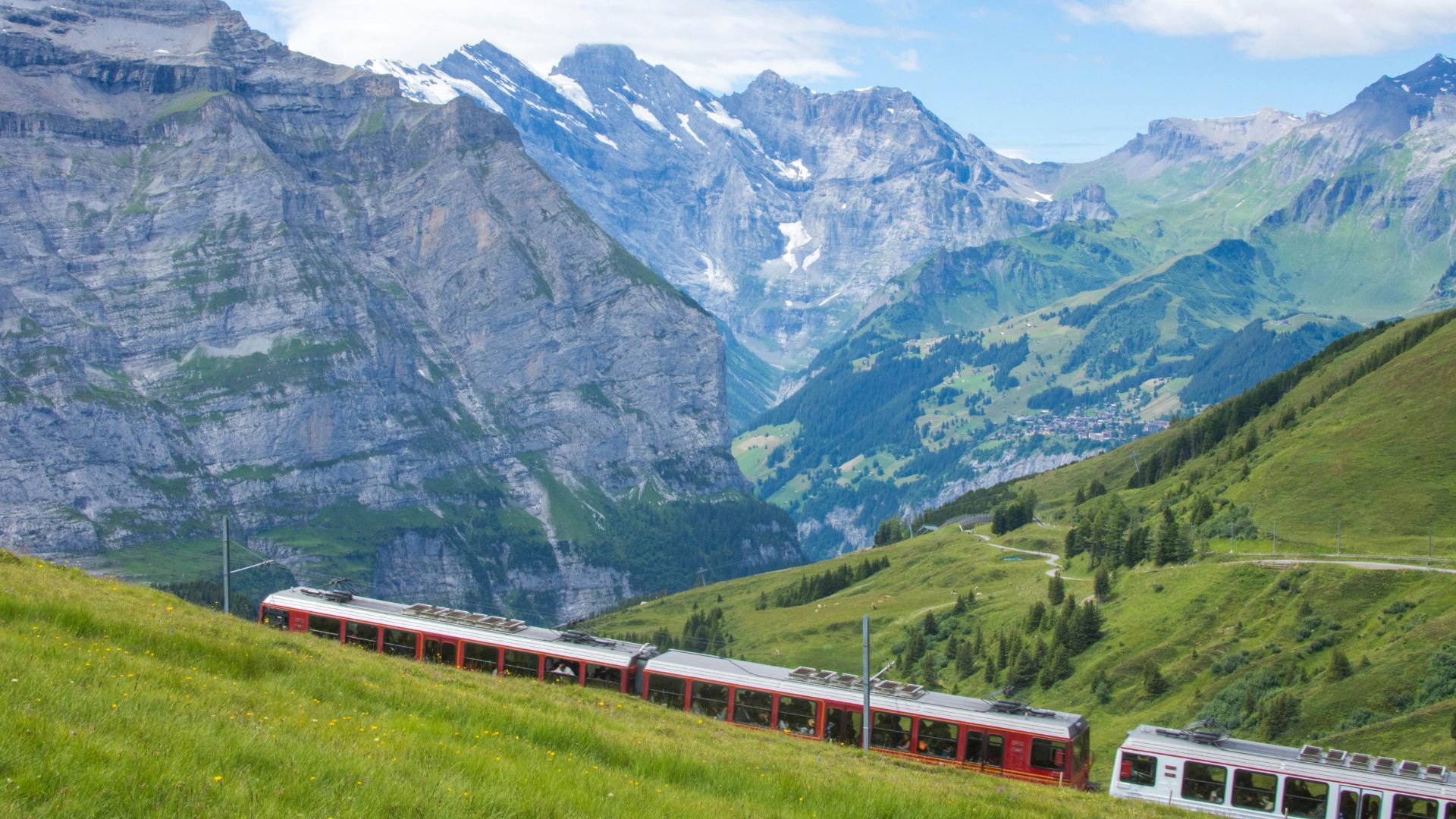 A red and white cogwheel train ascends a steep, bright green mountain slope in the Swiss Alps, with a towering, snow-capped mountain face in the background under a blue sky with white clouds.