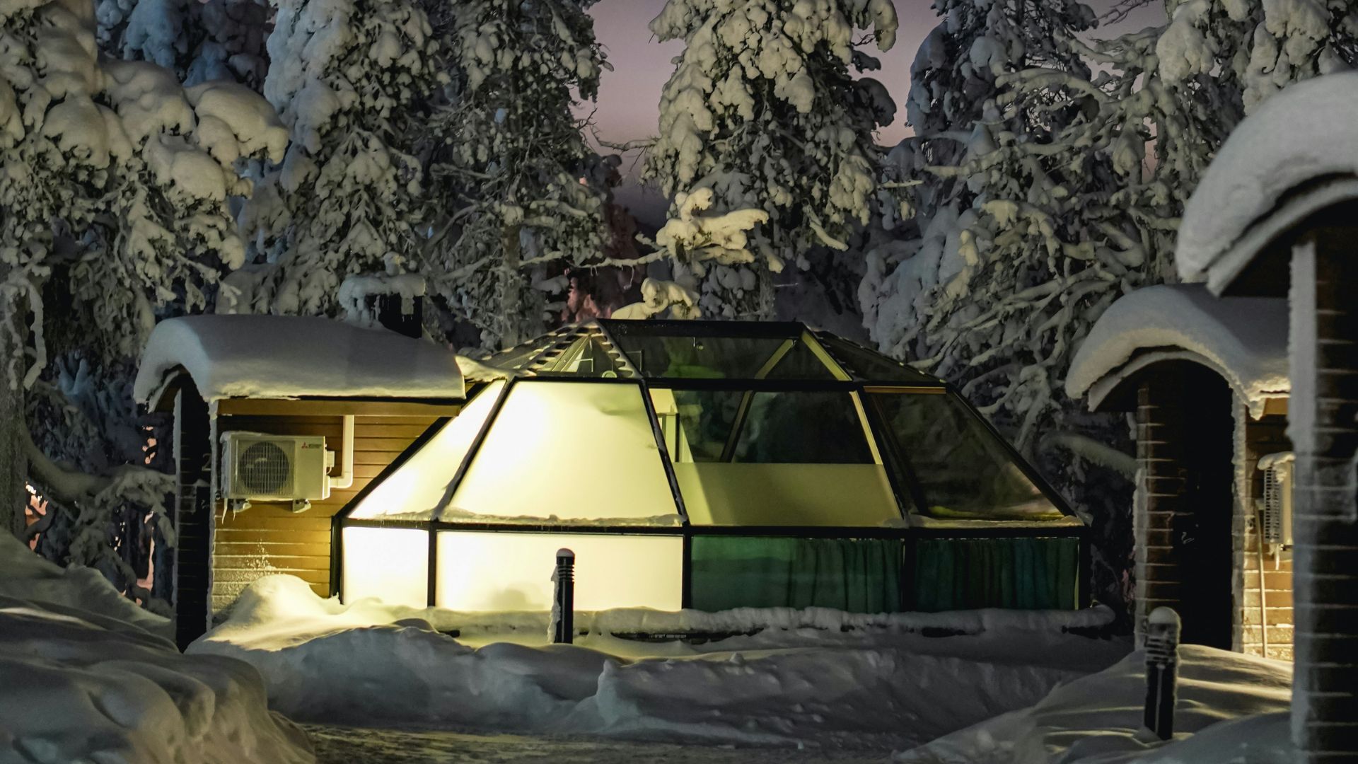 A dome-shaped glass igloo is illuminated from within at night, surrounded by deep snow and snow-covered pine trees in a quiet arctic resort setting.