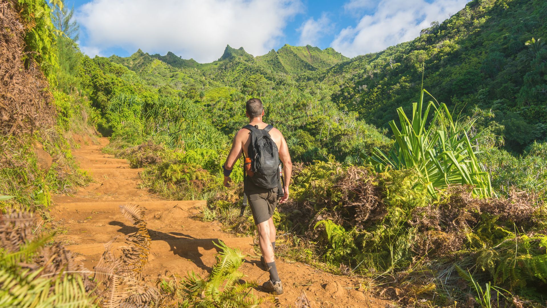A man in a tank top and backpack hikes up a steep, reddish-dirt trail in Kauai, Hawaii, surrounded by lush green tropical plants and dramatic, pointed mountain peaks under a blue sky.
