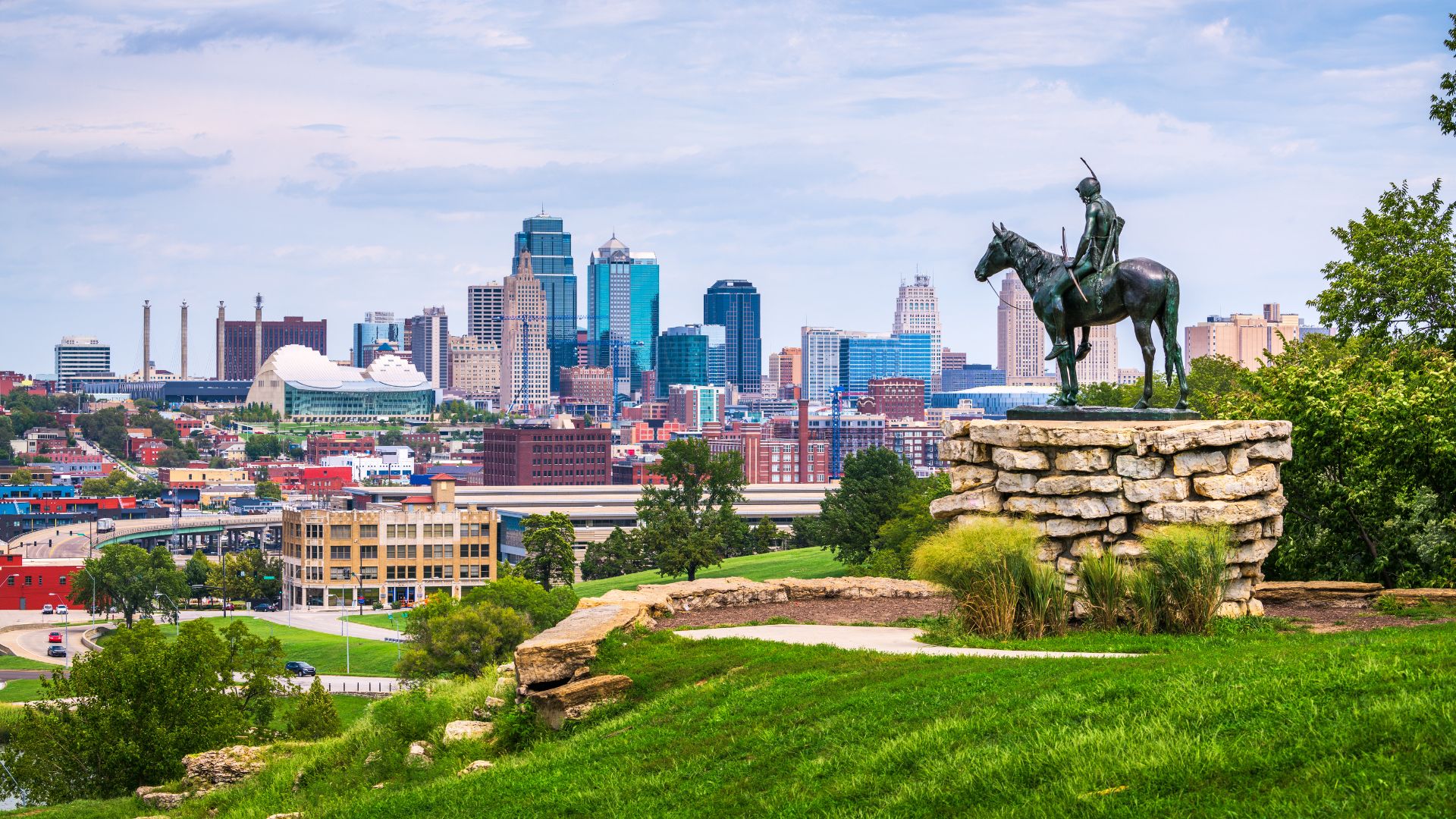 An iconic bronze statue of a Native American on horseback, known as 'The Scout', stands on a rocky pedestal on a grassy hill overlooking the extensive, modern skyline of downtown Kansas City.