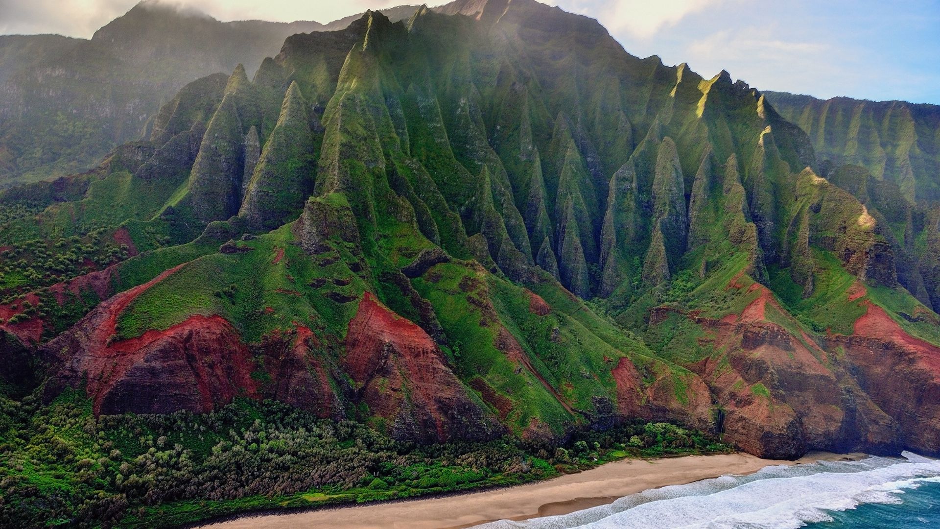 An aerial photograph of the Nā Pali Coast in Kauaʻi, Hawaii, showing massive, rugged green and red-soiled cliffs with razor-sharp ridges towering over a sandy beach and the blue ocean.
