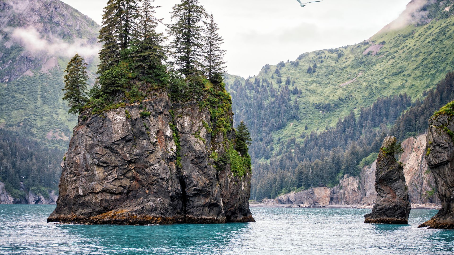 A tall, rugged rock pillar topped with evergreen trees stands in a bay with blue water, surrounded by forested mountain cliffs under an overcast sky.