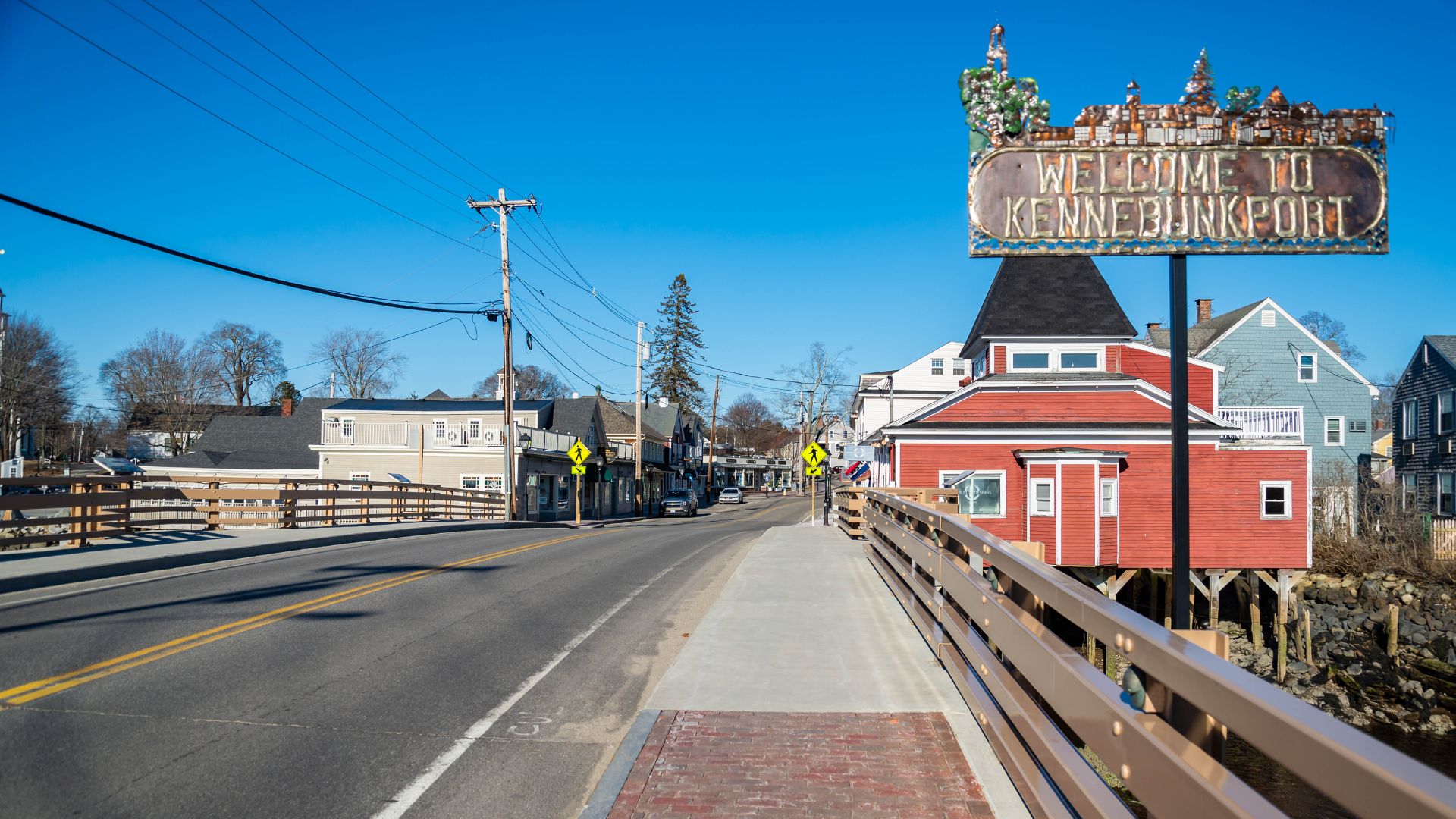A street and bridge leading into the village of Kennebunkport, Maine, with shops and houses along the waterfront under a clear blue sky.