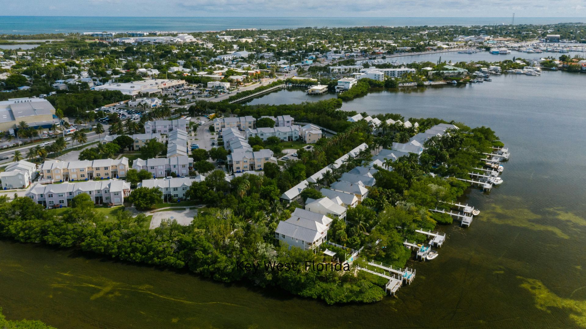 An aerial photograph of a coastal residential community in Key West, Florida, featuring white townhomes, lush green mangrove vegetation, numerous boat docks, and the blue ocean in the background.