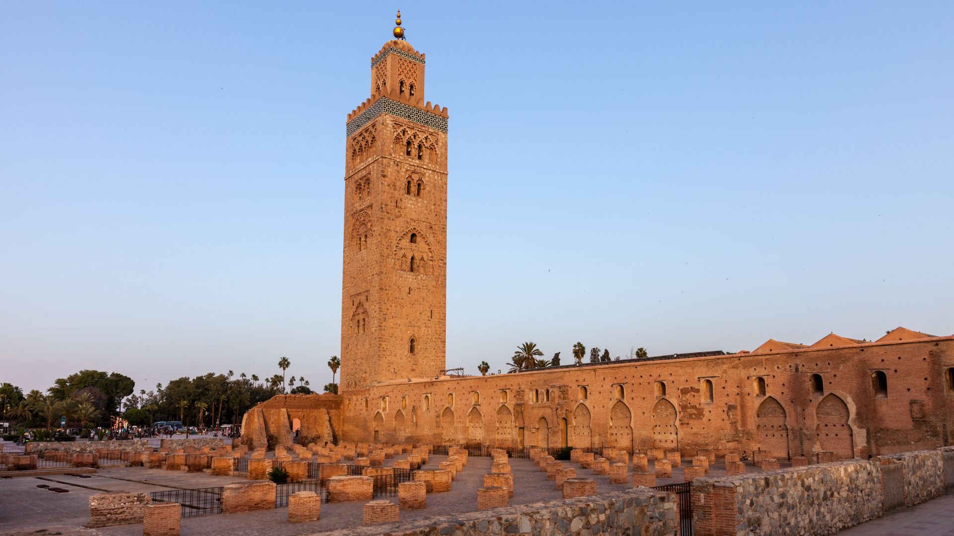 A tall, square sandstone minaret of the Koutoubia Mosque in Marrakesh, Morocco, with the mosque structure and ancient ruins in the foreground under a clear sky.