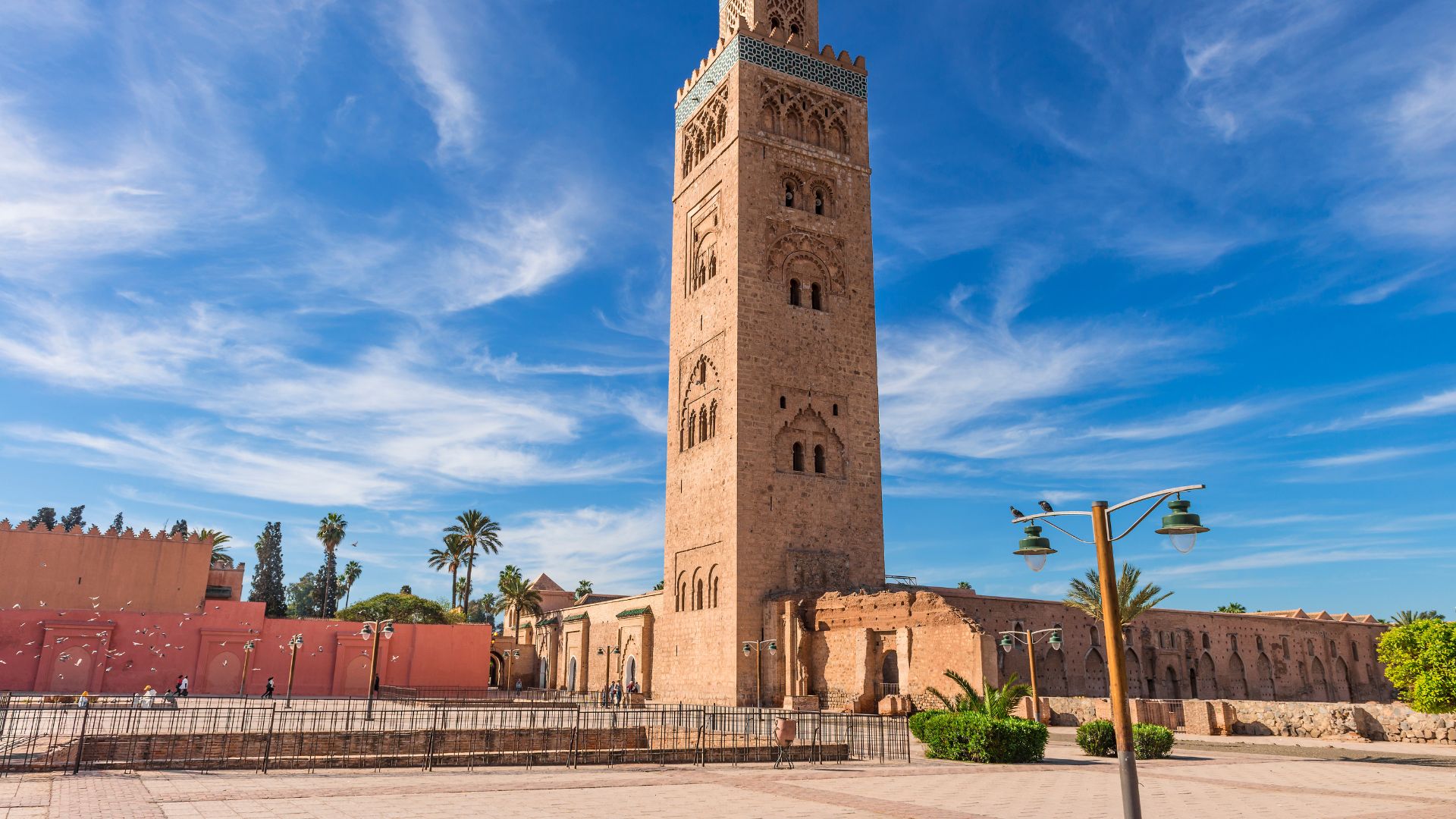 A large, square minaret of the Koutoubia Mosque in Marrakesh, Morocco, under a blue sky with white clouds.