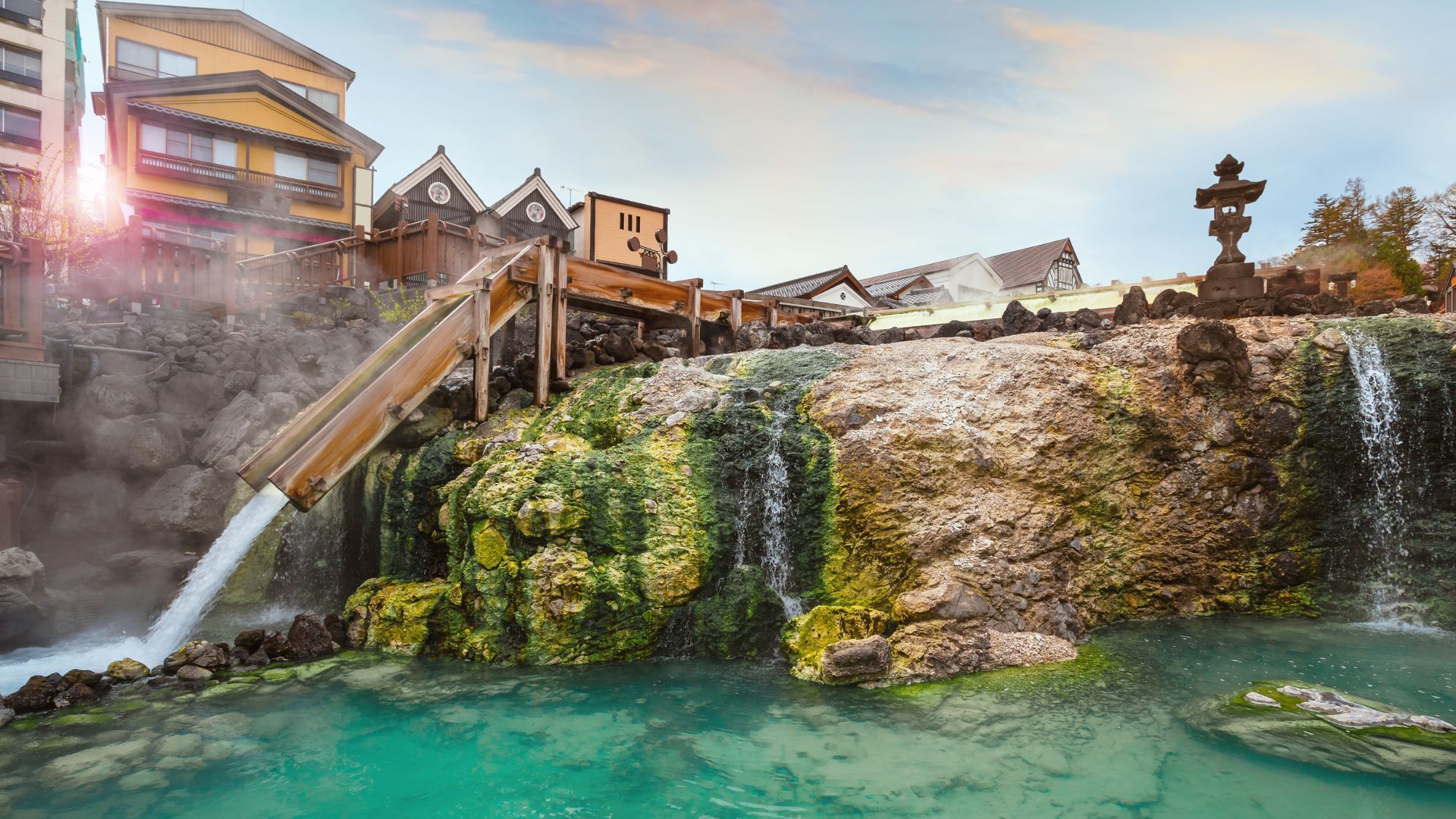 A central hot spring system in Kusatsu Onsen, Japan, featuring milky blue water, steam, wooden troughs, and traditional buildings on a hillside.