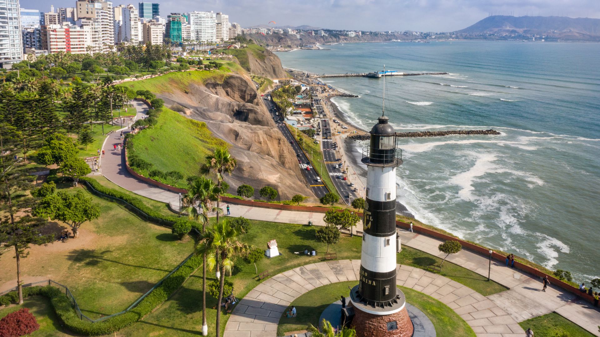 A black and white striped lighthouse in a park on cliffs overlooking the ocean and a city skyline.