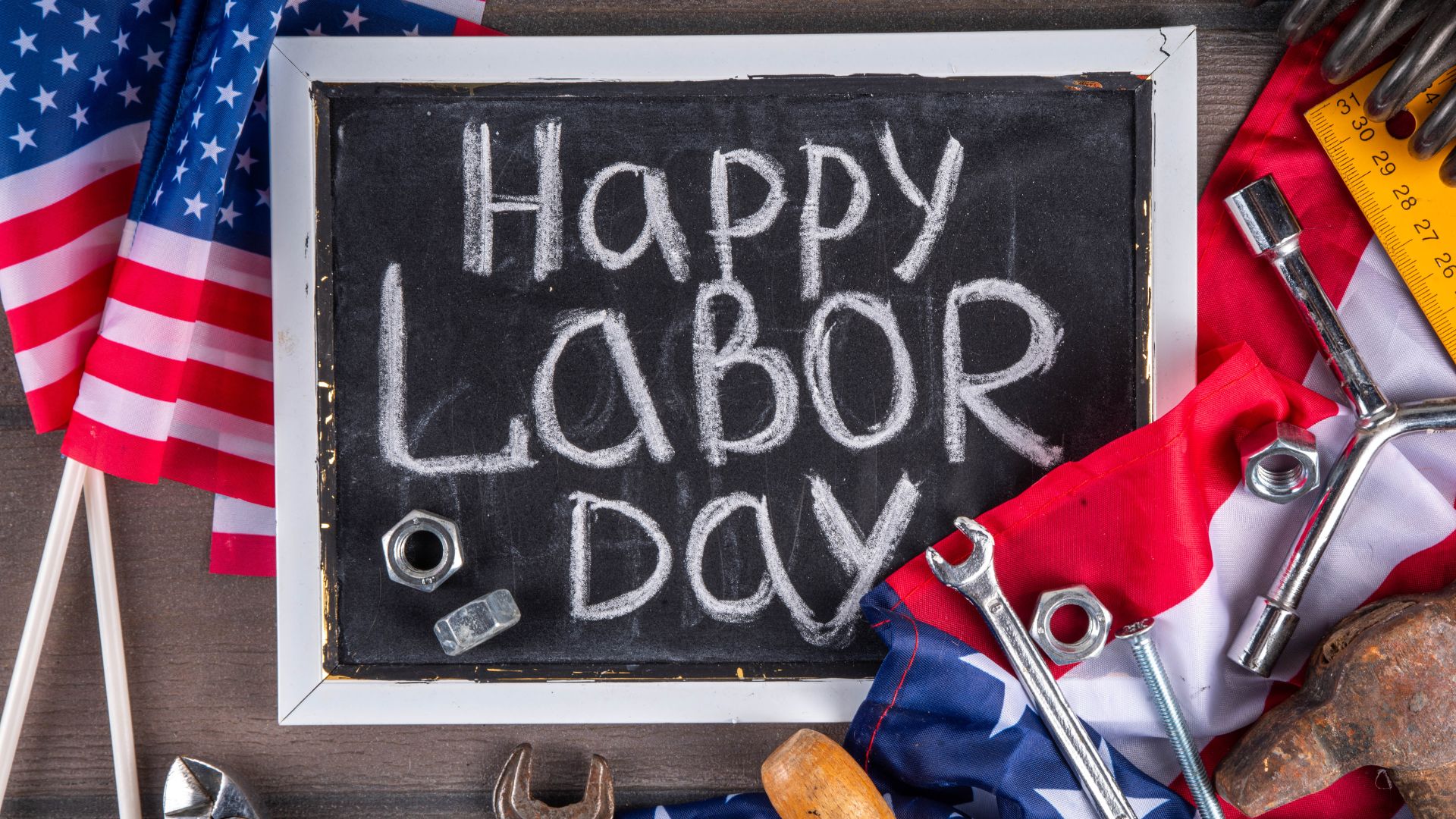 A chalkboard sign with "Happy LABOR Day" written in chalk, surrounded by American flags and various construction tools like wrenches and nuts.