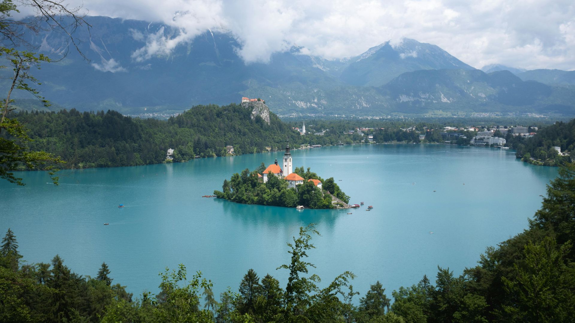 An elevated view of the turquoise Lake Bled, with a church on an island in the center and a castle on a cliff overlooking the lake, surrounded by mountains.