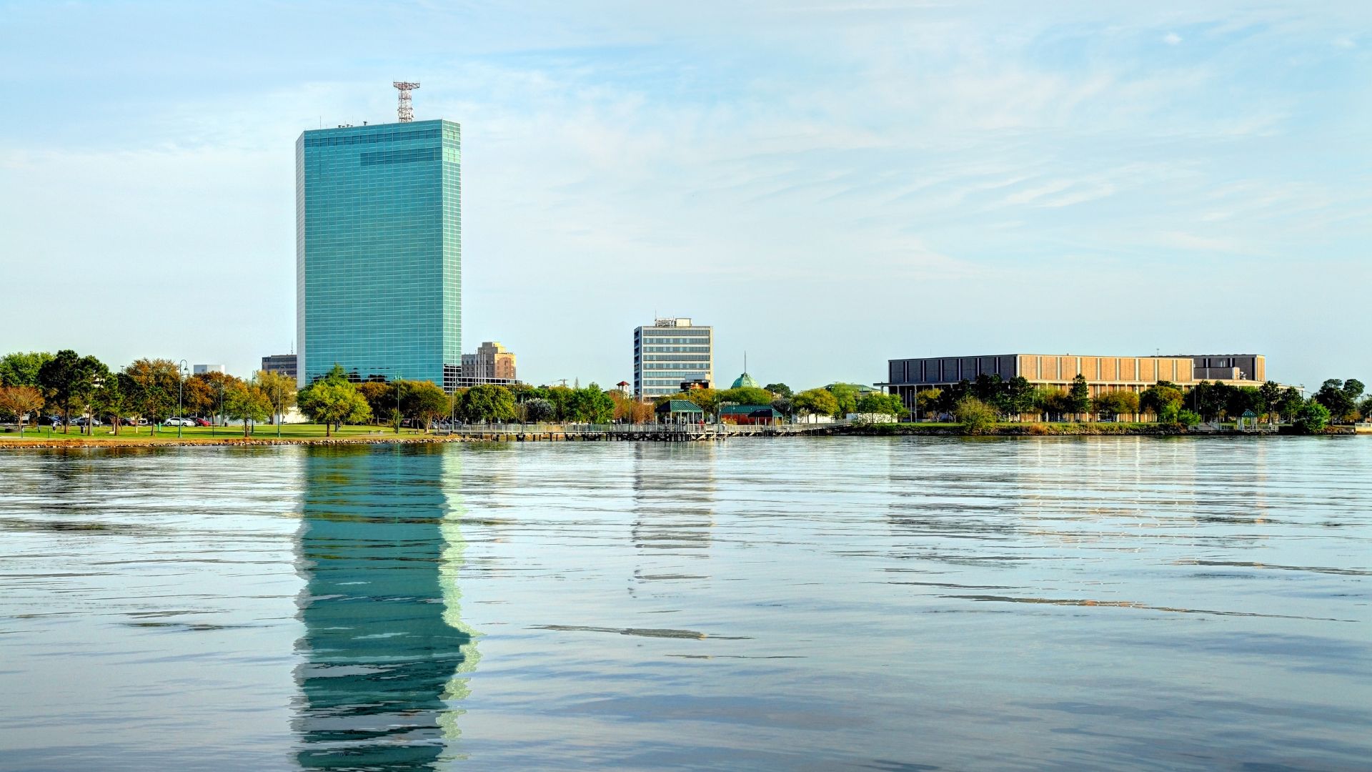 A daytime view of a city skyline with a tall, teal-glass skyscraper and a large civic center building along a waterfront.