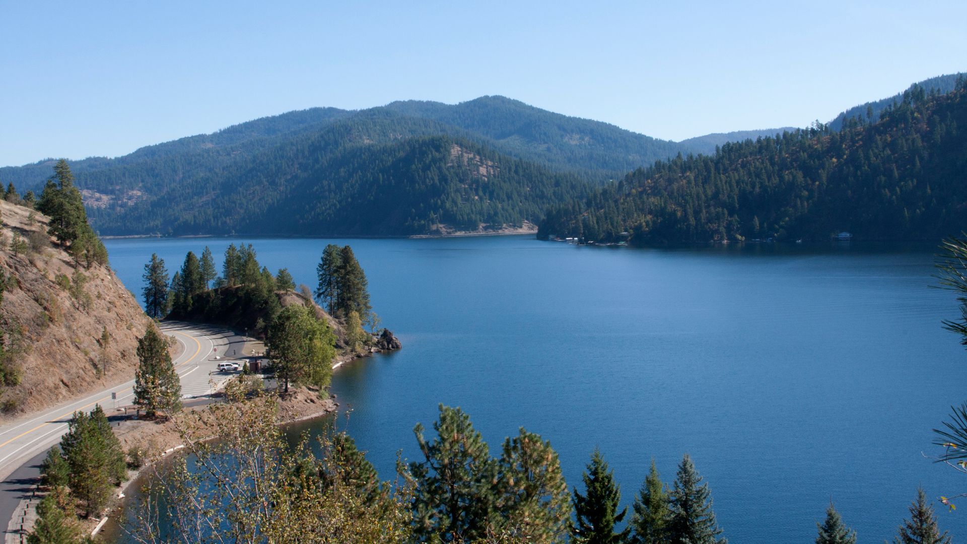 A wide, blue mountain lake surrounded by steep, densely forested hillsides under a clear sky, with a winding road visible along the near shore.