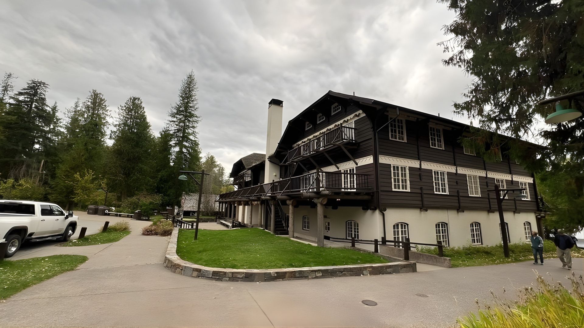 A large, historic lodge building with a white lower level and dark wood siding upper levels and balconies, set amidst pine trees next to a paved pathway.