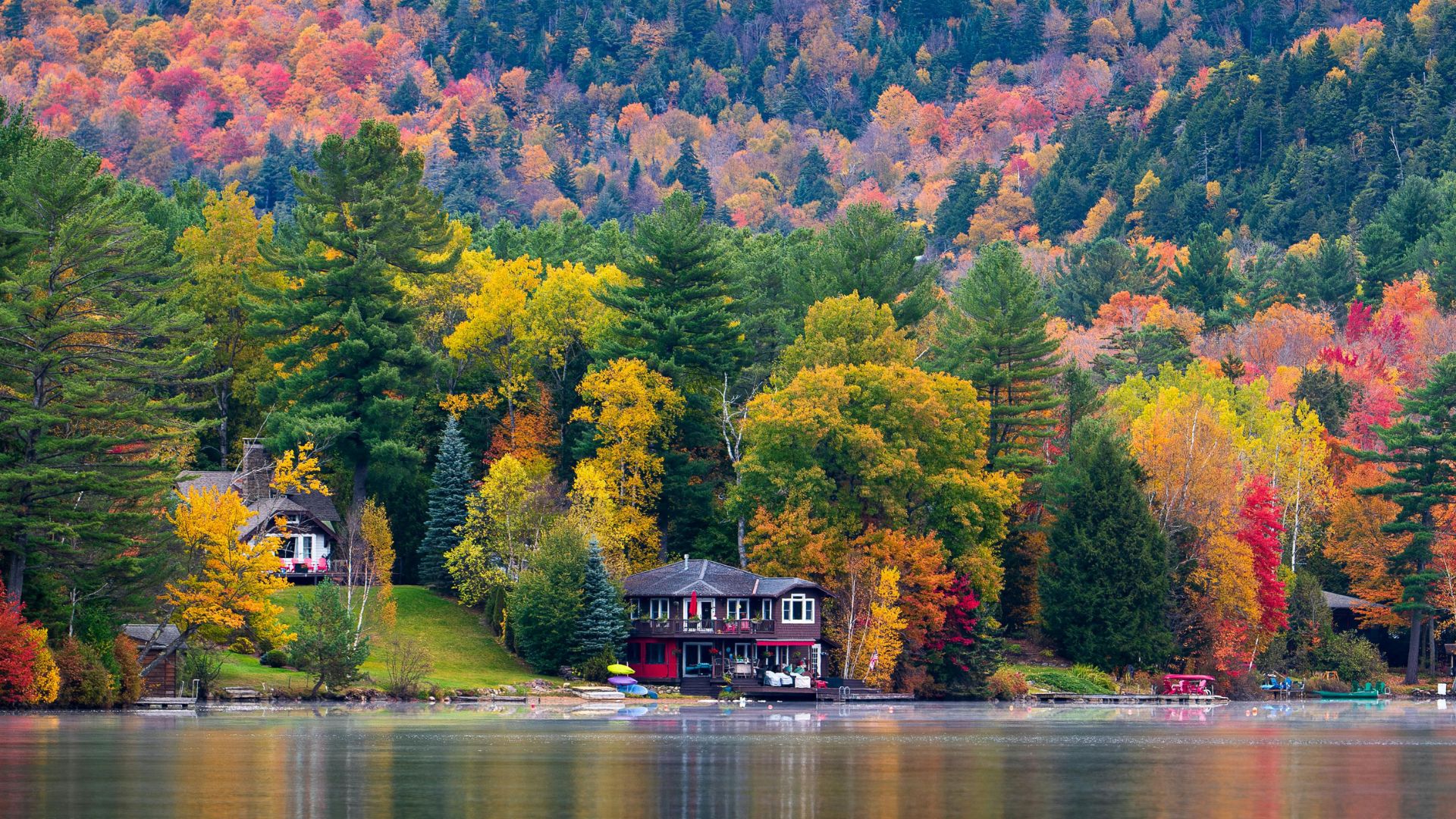 A scenic view across a calm lake in the Adirondack Mountains, showing a dense forest on the opposite shore with a mix of green, yellow, and red fall foliage. A mountain with a visible ski slope rises in the distance.