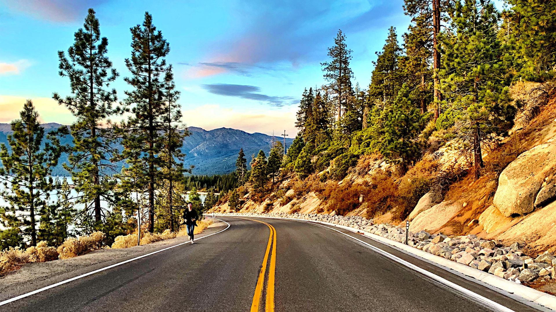 A person runs along a curved mountain highway next to the deep blue water of Lake Tahoe, surrounded by evergreen forests and mountains under a colorful sky at sunset.