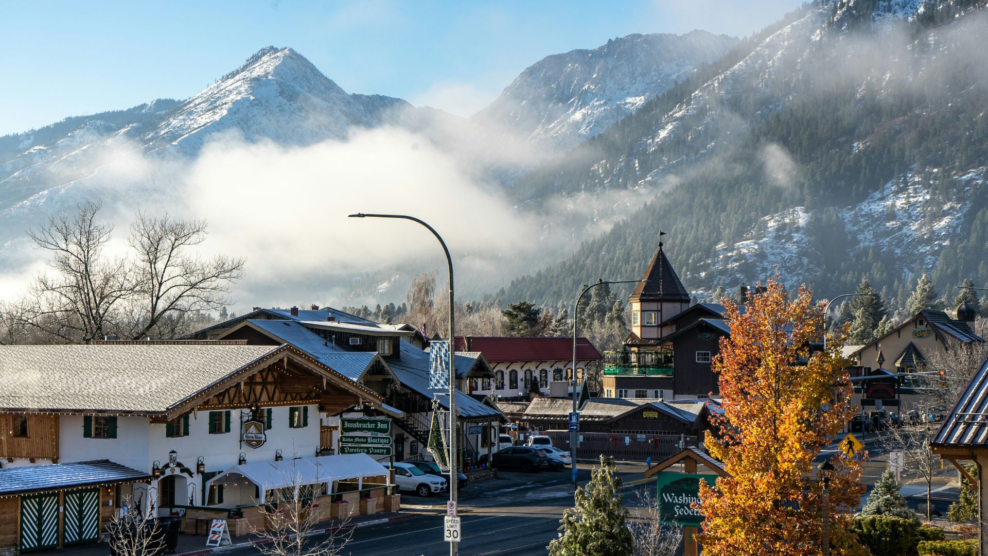 A panoramic view of the Bavarian-themed town of Leavenworth, Washington, featuring buildings with half-timbering and gabled roofs in the foreground, backed by a coniferous forest and snow-capped mountains under a bright, partly cloudy sky.