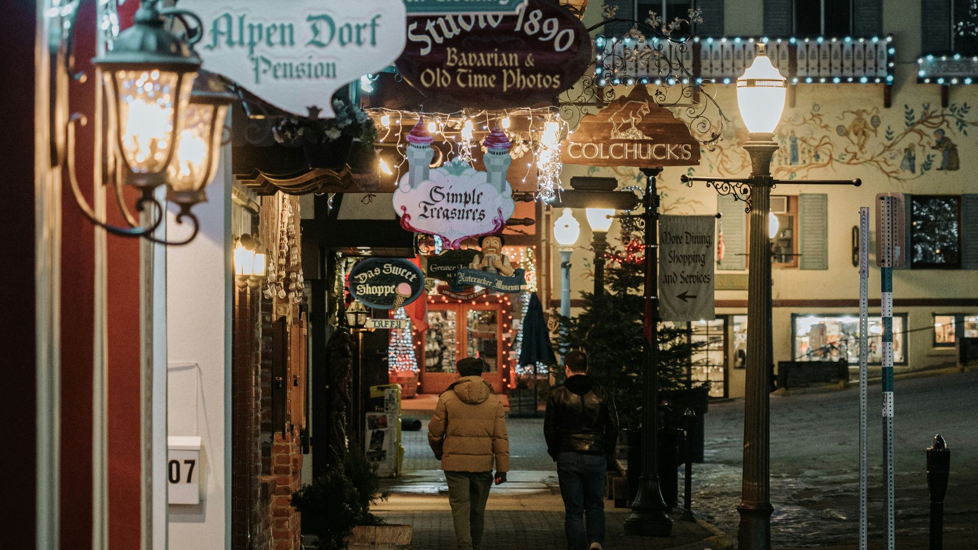 A snowy street in a Bavarian-themed village at night with holiday lights and people walking.