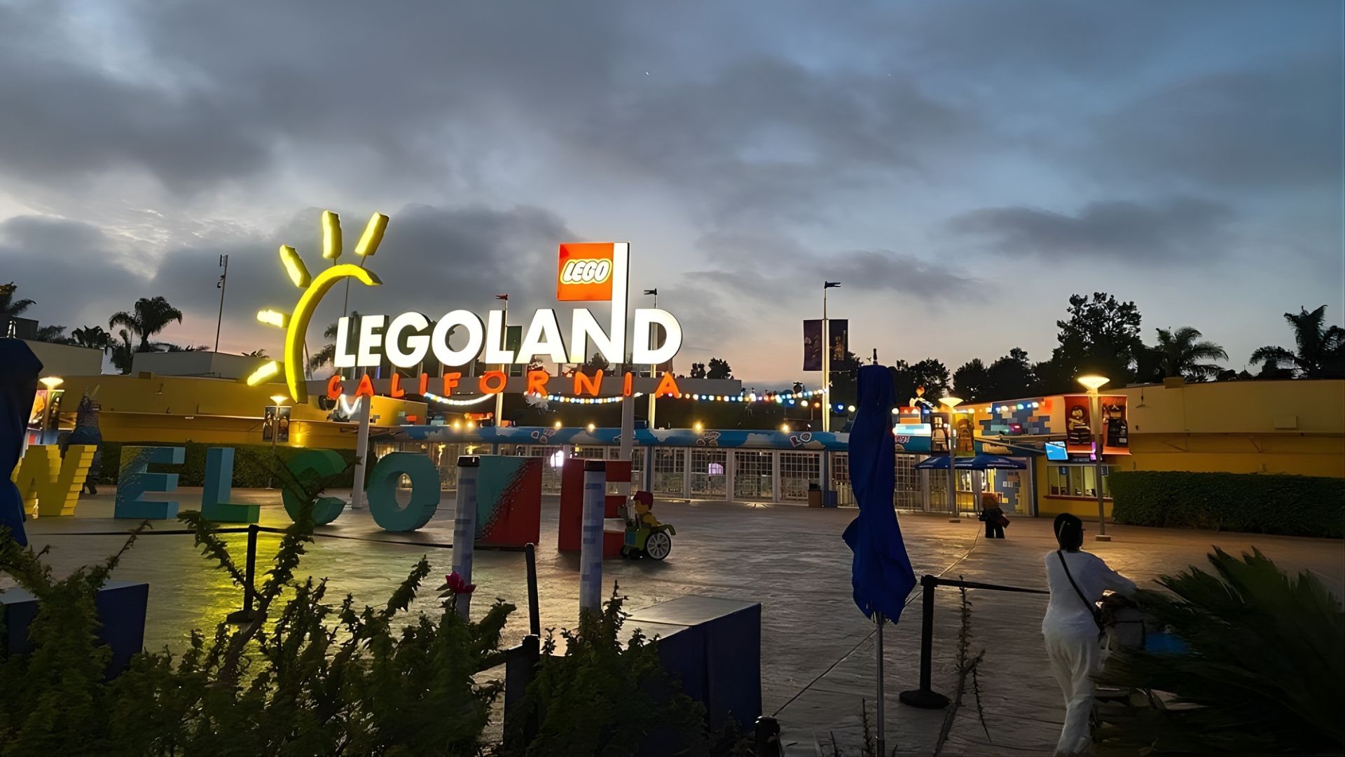 The entrance to the LEGOLAND California theme park at dusk, featuring large, illuminated yellow 'LEGOLAND' signage and a 'WELCOME' sign, with some people and park facilities visible.