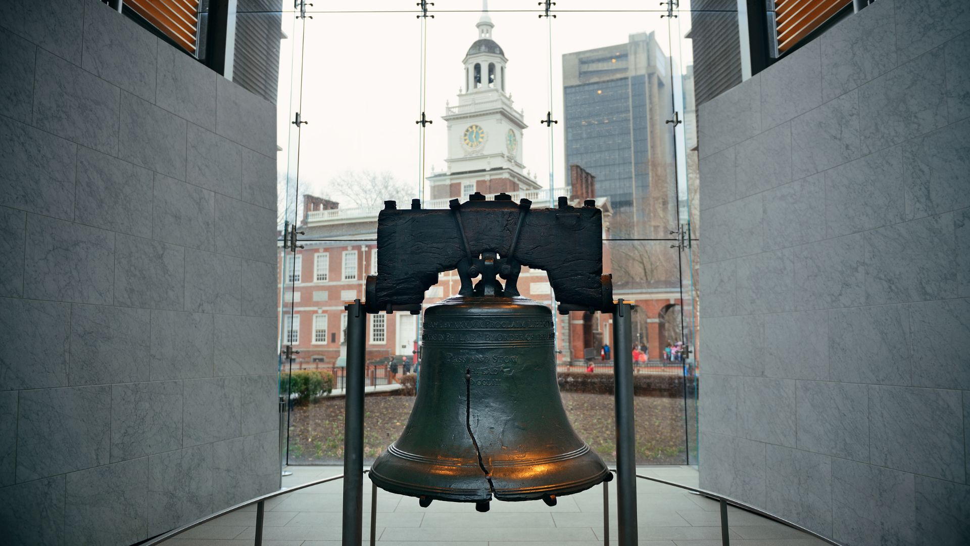 The cracked Liberty Bell displayed inside a modern glass pavilion, with the historic, red-brick Independence Hall visible through the window in the background.