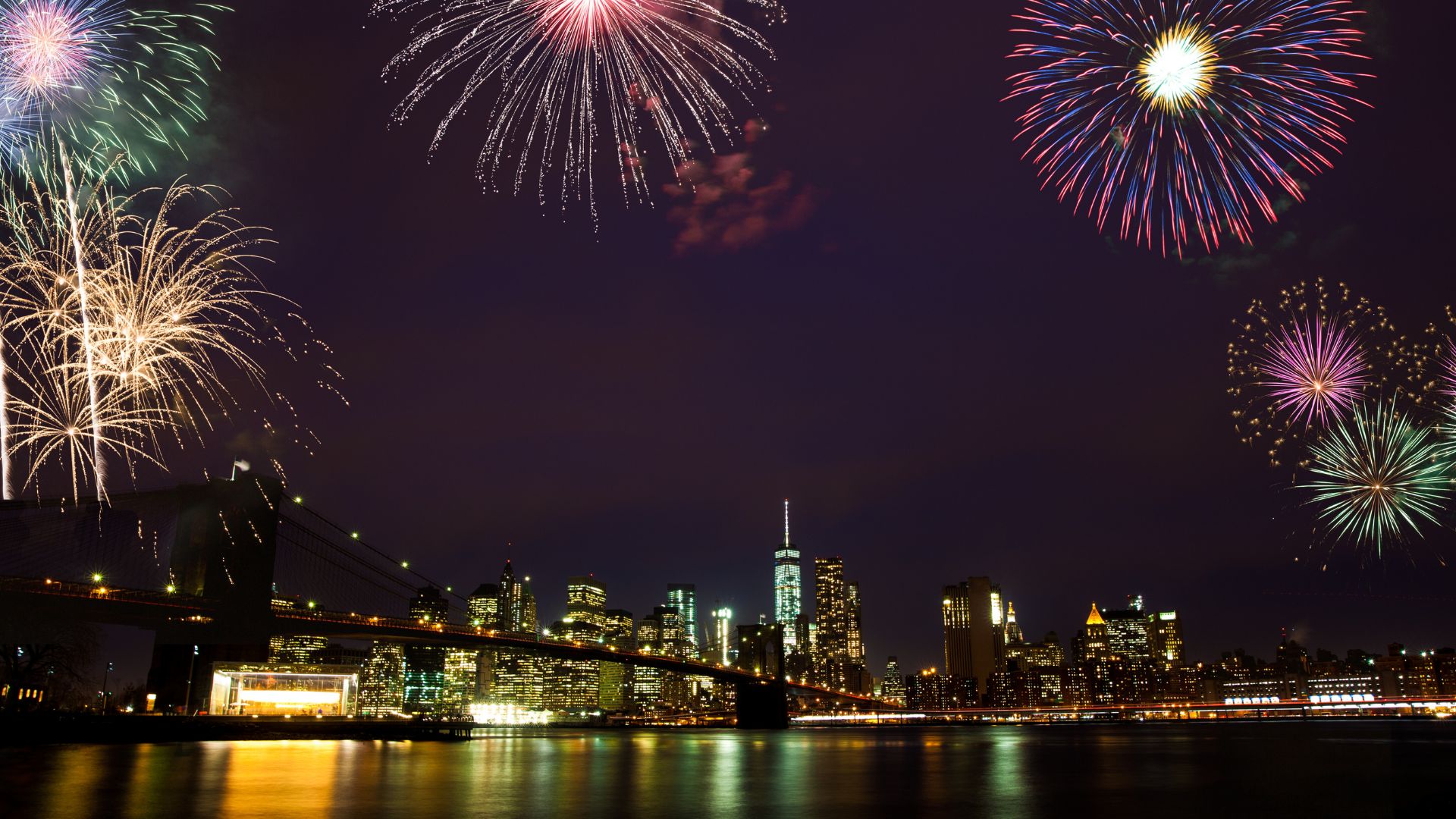 Nighttime panoramic view of the Manhattan skyline and the Brooklyn Bridge with colorful fireworks exploding above.