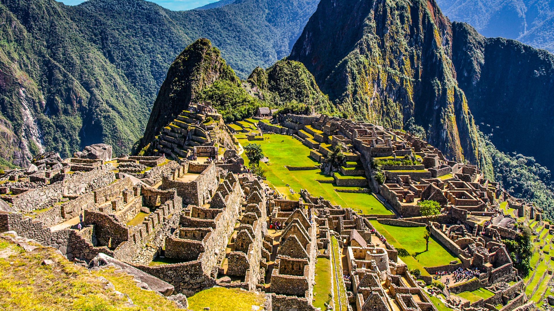 A panoramic view of the ancient Inca citadel of Machu Picchu, featuring stone buildings, agricultural terraces, and lush green mountains in the background.