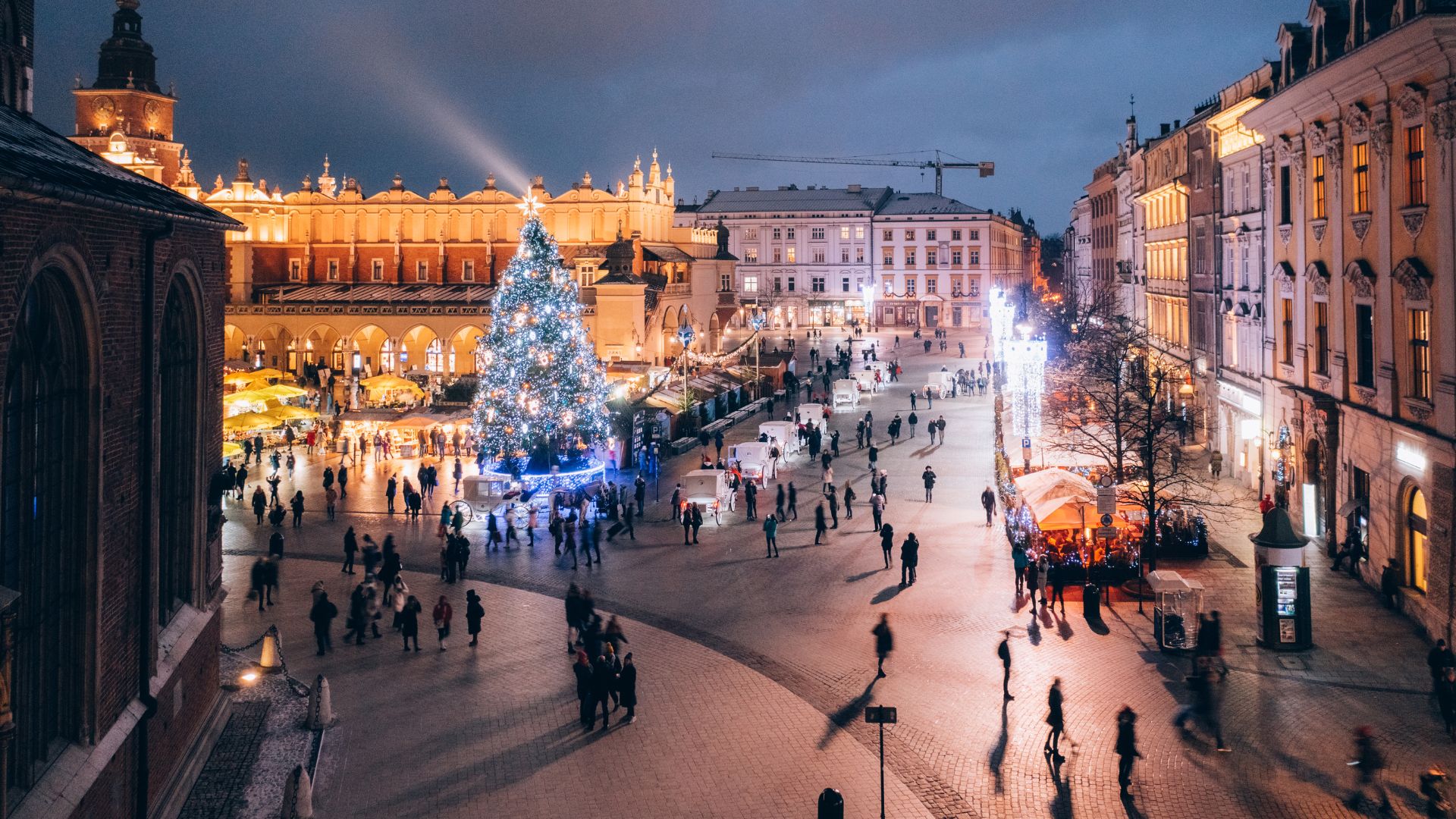 A festive, evening photograph of the bustling Main Market Square in Kraków, Poland, featuring a large, illuminated Christmas tree and market stalls in the foreground, with the historic Cloth Hall and its tower prominently lit in the background.