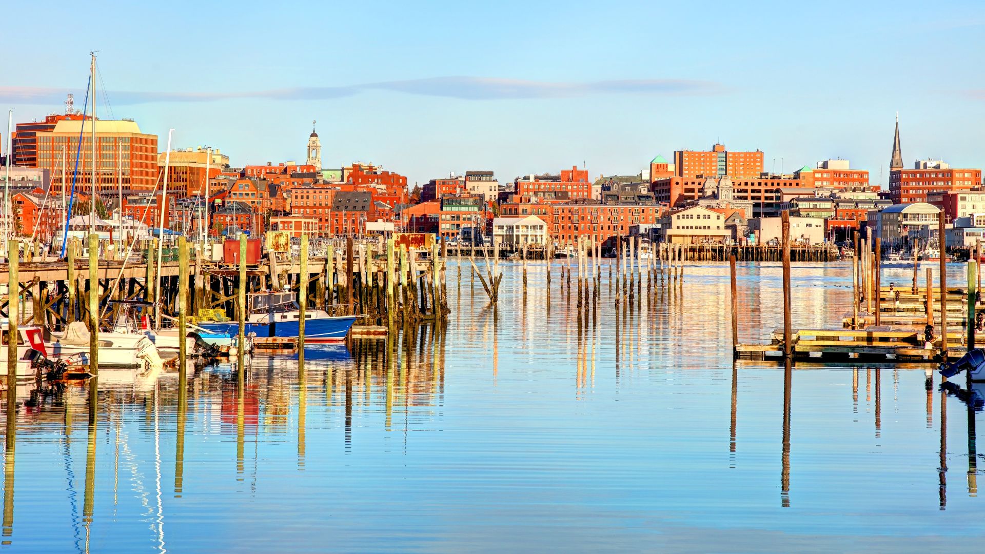 A panoramic view of the Portland, Maine, waterfront, featuring numerous boats docked in a busy harbor with tall, wooden pilings, reflecting the historic, red-brick cityscape and spires on the shore under a bright blue sky.