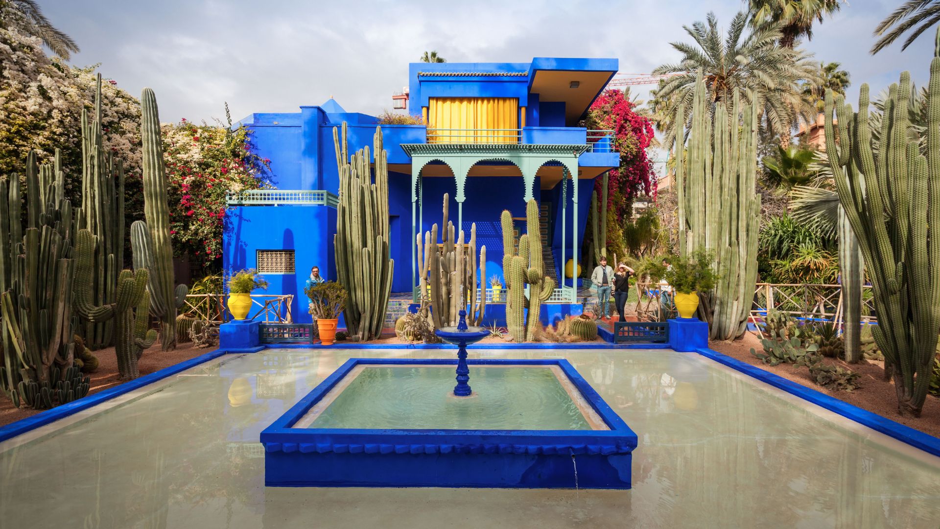 A vibrant cobalt blue building surrounded by tall, sculptural cacti and lush vegetation, with a small reflective pond and fountain in the foreground.
