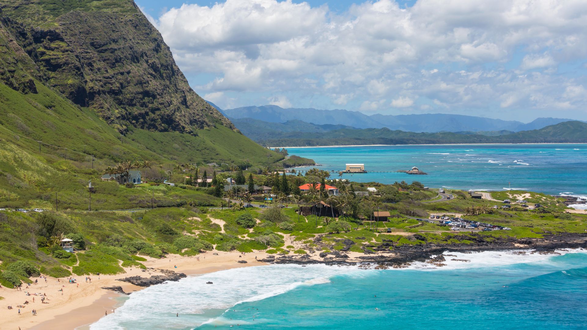 An elevated, panoramic view of a beautiful Hawaiian coastline featuring a sandy beach with crashing turquoise waves, a steep green mountain on the left, and distant islands in a bay under a bright, cloudy sky.